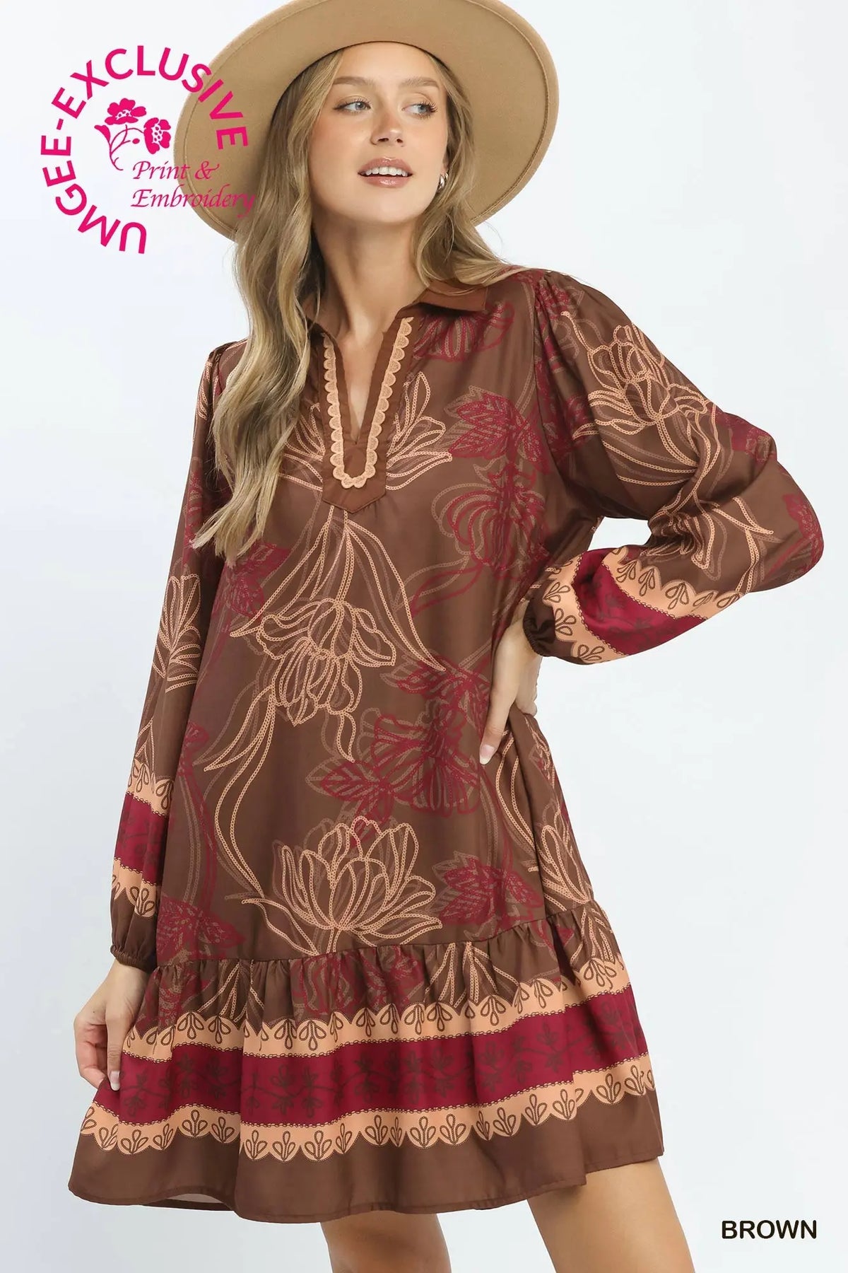 A woman modeling a brown long-sleeve bohemian-style mini dress with a beige floral print and a deep red patterned hem, wearing a wide-brimmed tan hat against a white studio background.