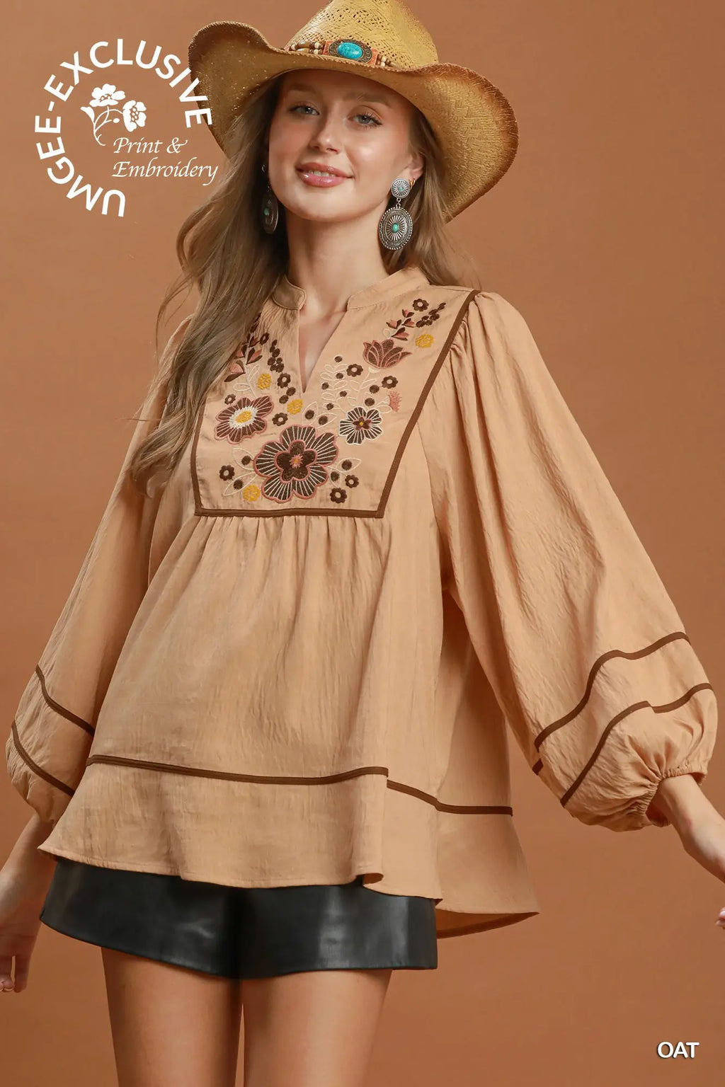 A woman wearing a tan bohemian-style blouse with floral embroidery on the chest, a matching straw hat, and black shorts, posed against a solid warm brown background. A circular logo for 'Unbe-E Exclusive Print & Embroidery' is visible in the top left, and 'OAT' is in the bottom right.