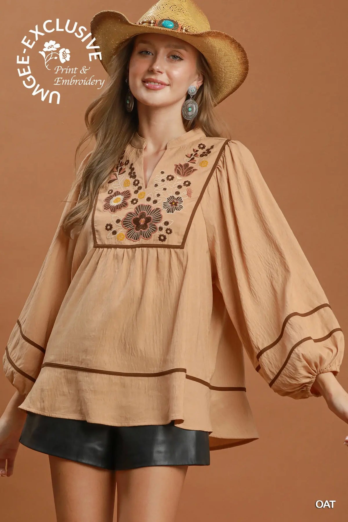 A woman wearing a tan bohemian-style blouse with floral embroidery on the chest, a matching straw hat, and black shorts, posed against a solid warm brown background. A circular logo for 'Unbe-E Exclusive Print & Embroidery' is visible in the top left, and 'OAT' is in the bottom right.