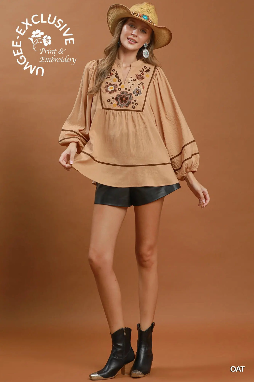 A woman models a tan long-sleeve blouse with floral embroidery on the chest, paired with black shorts and black ankle boots, against a solid brown studio background. A 'UGLEE EXCLUSIVE Print & Embroidery' logo is visible in the top left, and 'OAT' is in the bottom right.