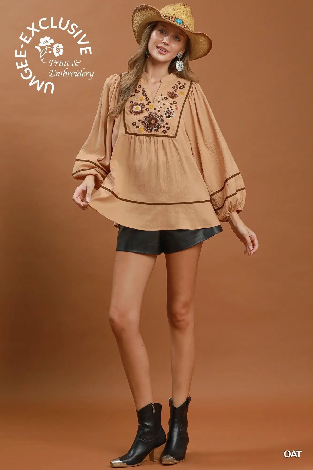 A woman models a tan long-sleeve blouse with floral embroidery on the chest, paired with black shorts and black ankle boots, against a solid brown studio background. A 'UGLEE EXCLUSIVE Print & Embroidery' logo is visible in the top left, and 'OAT' is in the bottom right.