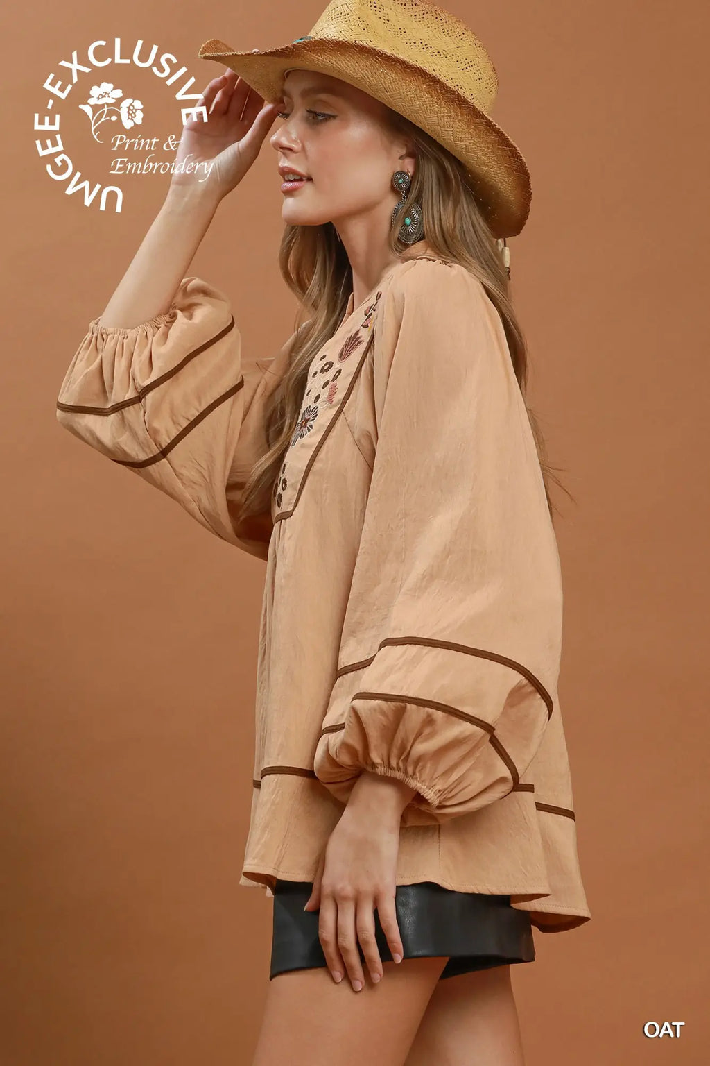 A woman wearing a beige bohemian-style blouse with brown trim, a straw hat, and black shorts, posed against a solid terracotta background. A white circular logo for 'UNIQUE EXCLUSIVE Print & Embroidery' is visible in the top left, and 'OAT' is printed in the bottom right.