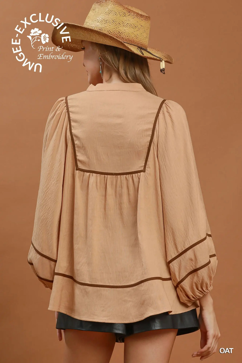A woman shown from the back wearing a beige long-sleeve blouse with dark brown trim and a straw hat, photographed against a warm tan studio background. A circular 'UNBE-EXCLUSIVE Print & Embroidery' watermark appears in the upper left, and 'OAT' is printed at the lower right.