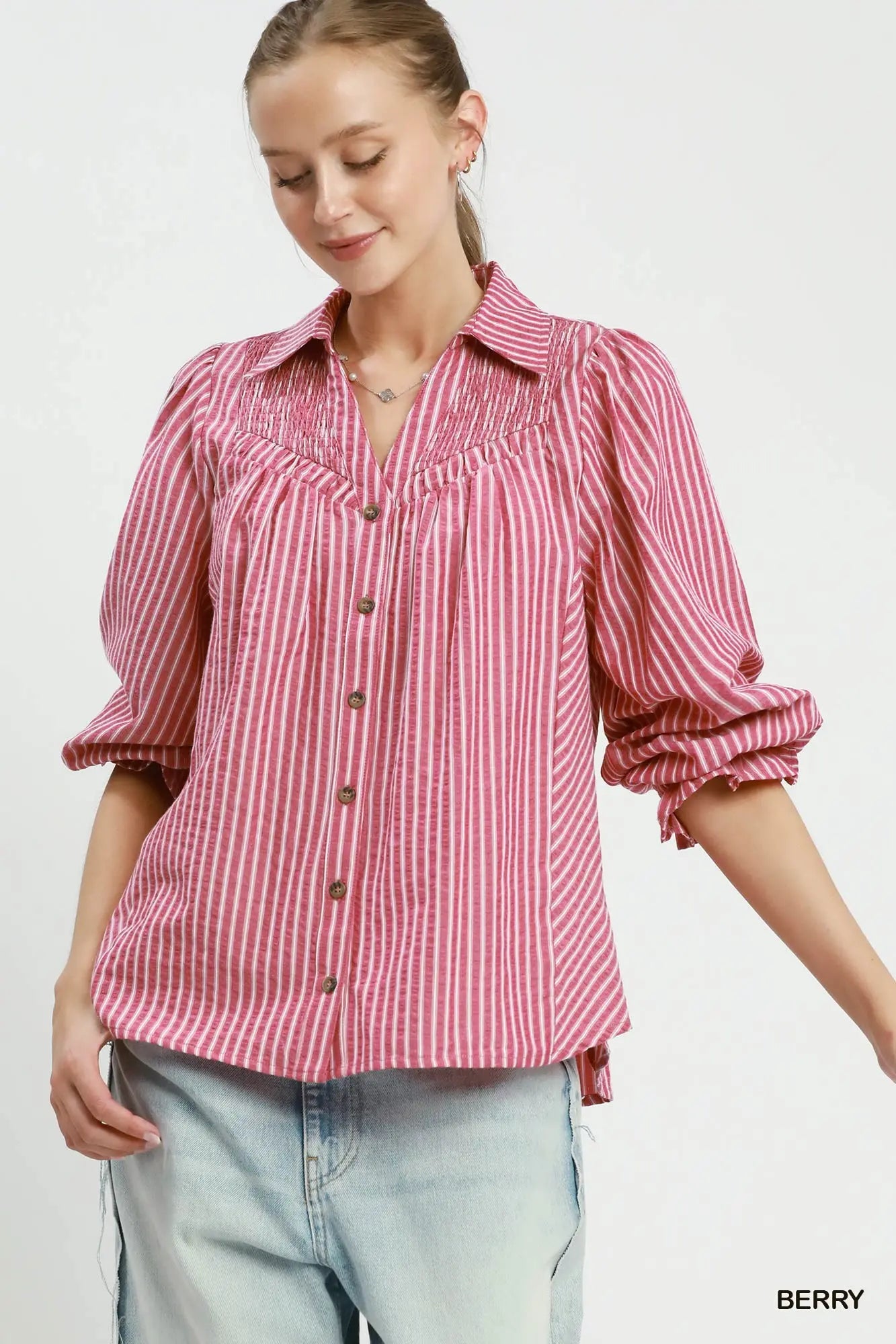 A woman wearing a pink and white vertically striped button-up blouse with ruffled detailing on the chest and rolled-up sleeves, paired with light blue jeans against a plain white background.