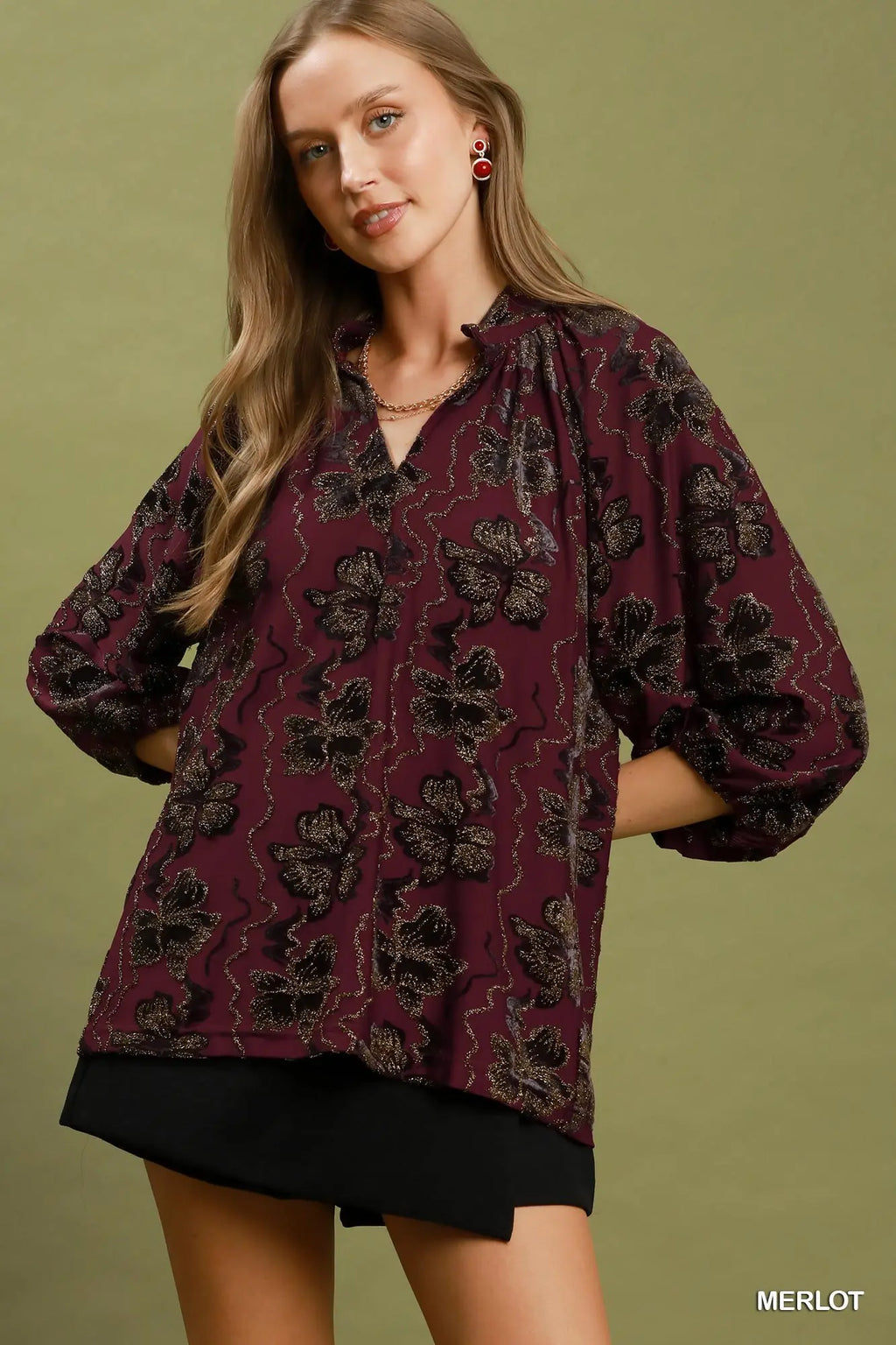 A woman modeling a loose-fitting long-sleeve blouse in a deep merlot color with a black floral pattern, paired with black shorts, against a solid olive green background.