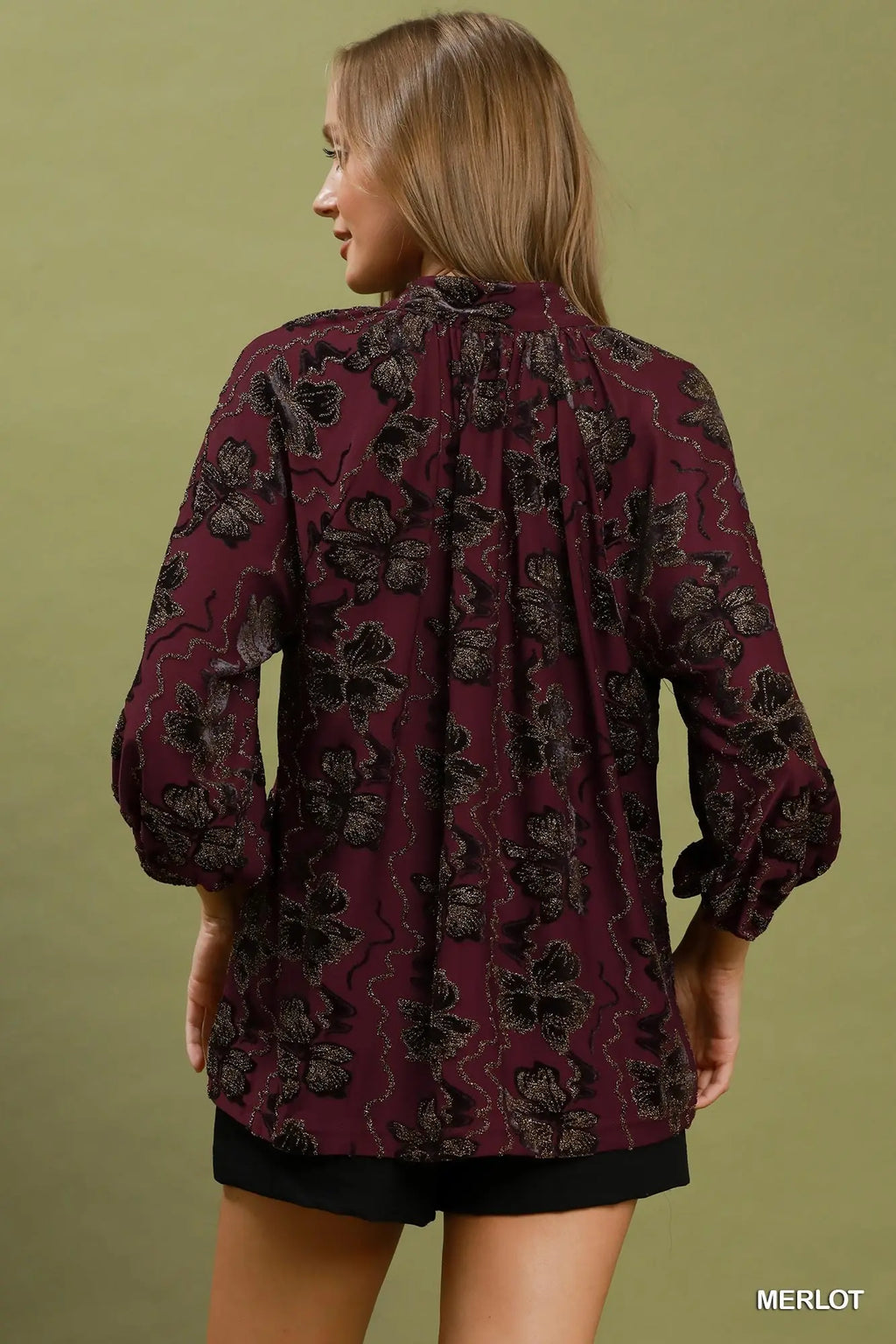 A woman shown from the back wearing a long-sleeve blouse in a deep burgundy or merlot color with a black floral pattern, paired with black shorts, against a solid olive green background.