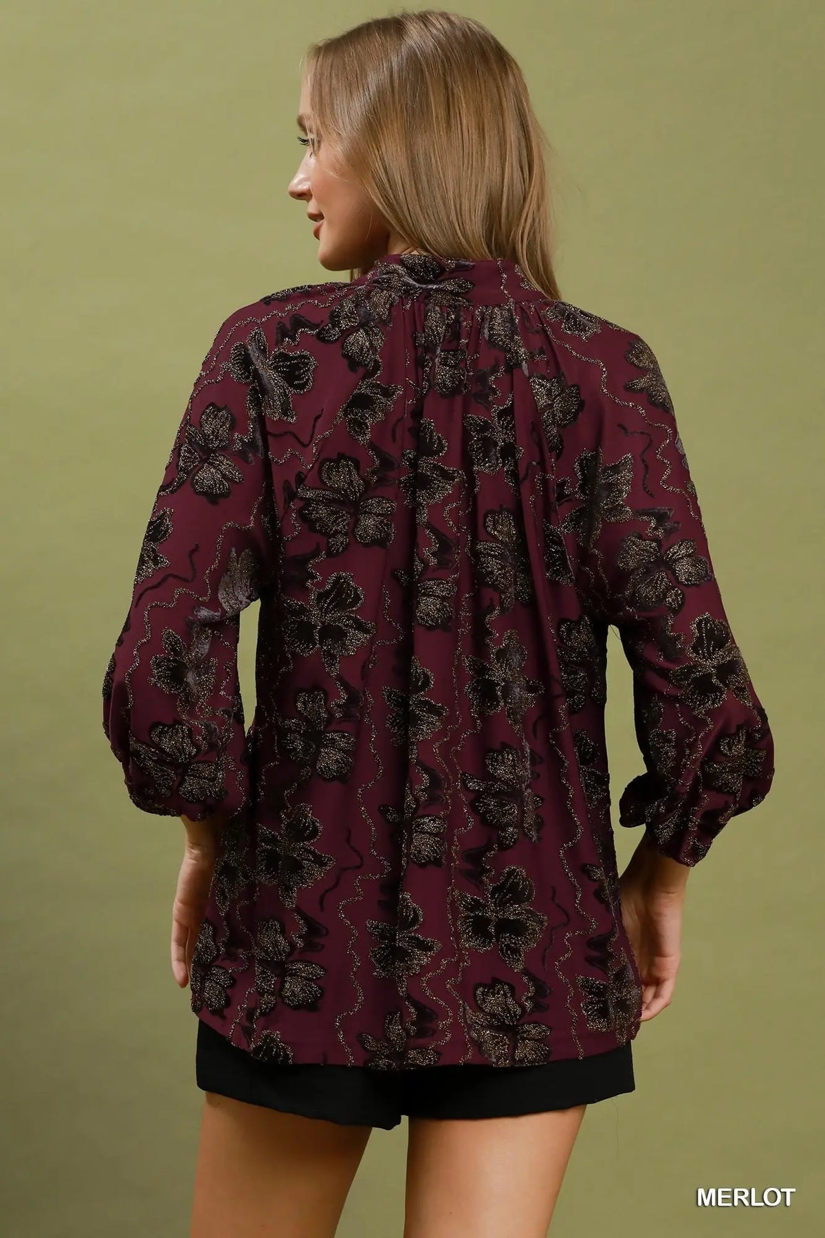 A woman shown from the back wearing a long-sleeve blouse in a deep burgundy or merlot color with a black floral pattern, paired with black shorts, against a solid olive green background.