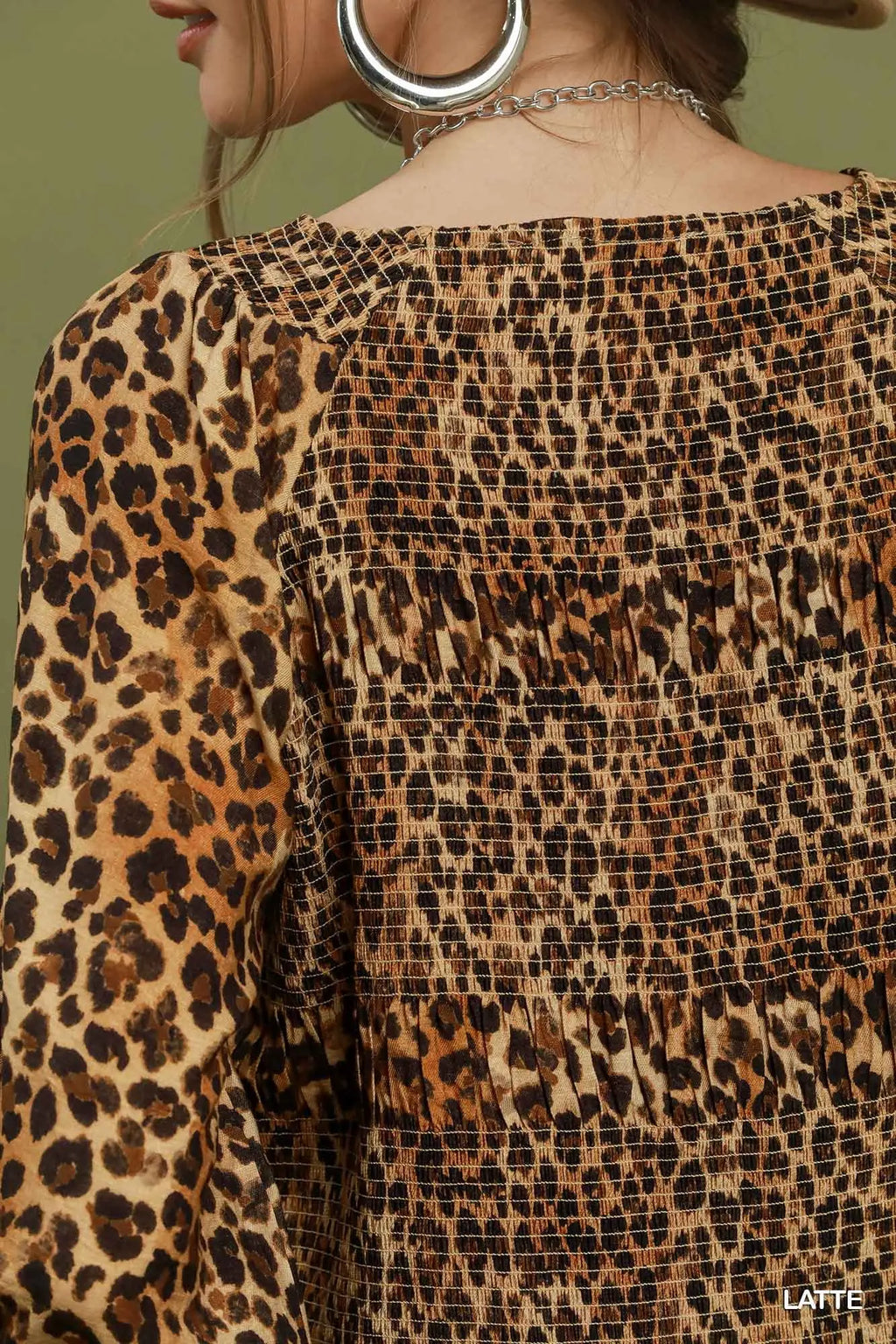 Close-up of a woman wearing a leopard print top with a smocked bodice and long sleeves, shown from the back against a muted olive background.