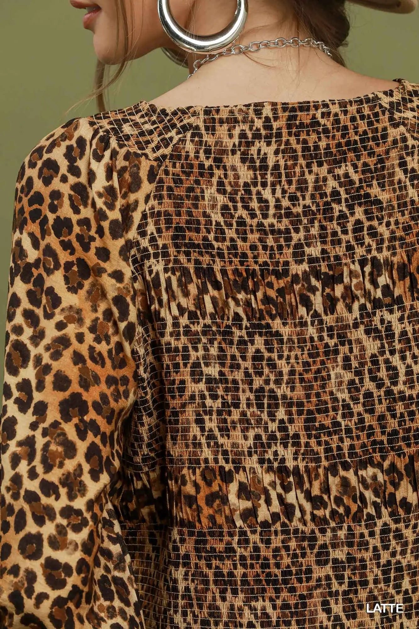 Close-up of a woman wearing a leopard print top with a smocked bodice and long sleeves, shown from the back against a muted olive background.