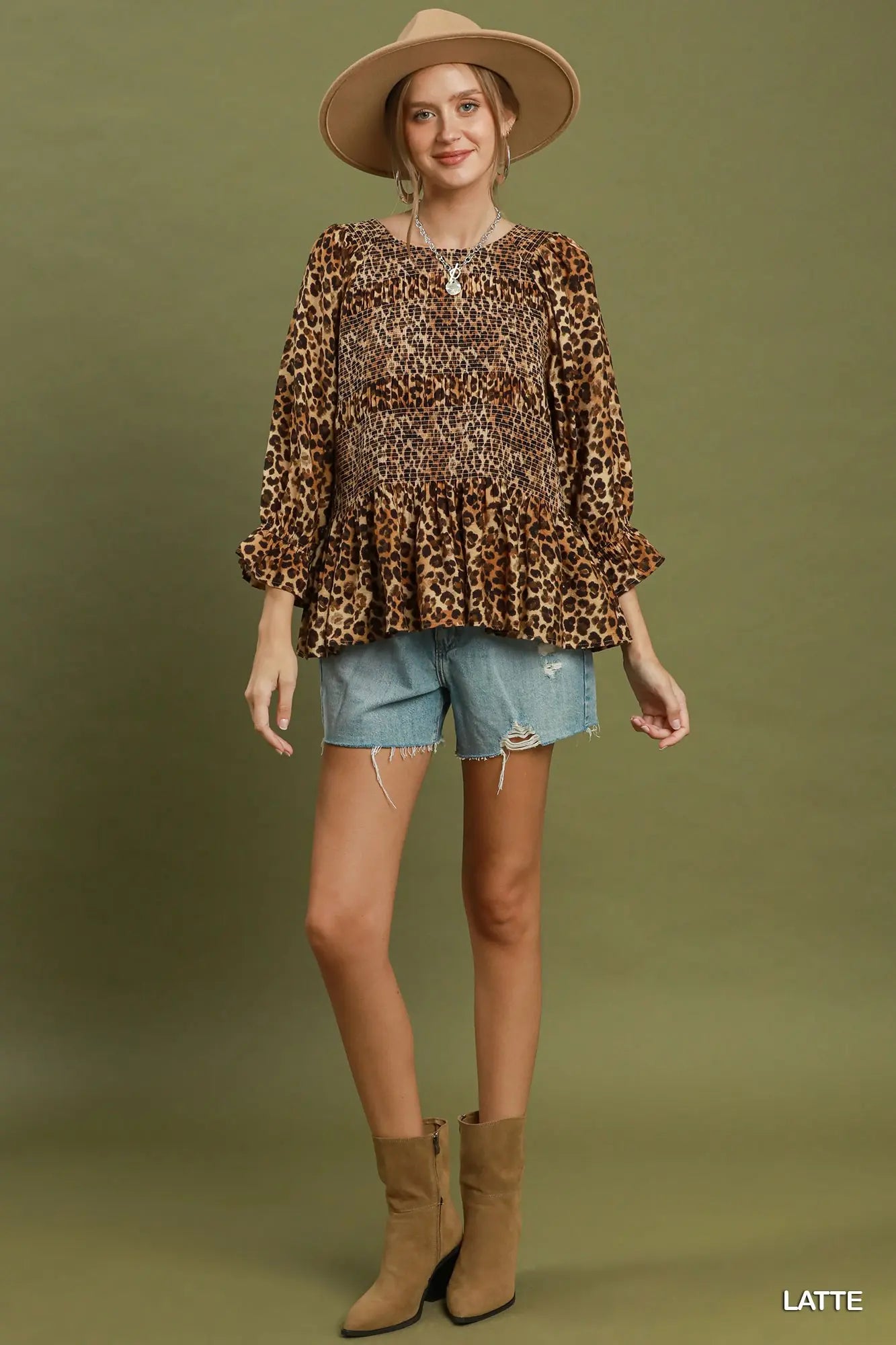 A woman modeling a long-sleeve leopard-print peplum top with light-wash denim shorts, tan ankle boots, and a wide-brim tan hat against an olive green studio background. The word 'LATTE' appears in the bottom right corner.