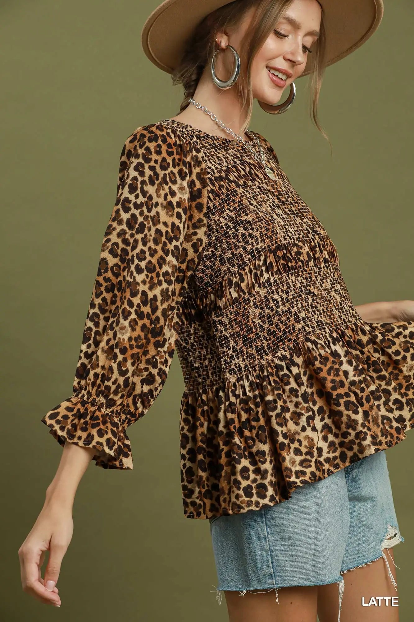 A woman wearing a leopard print blouse with three-quarter ruffled sleeves and a smocked bodice, paired with light blue jeans against a solid olive green background.