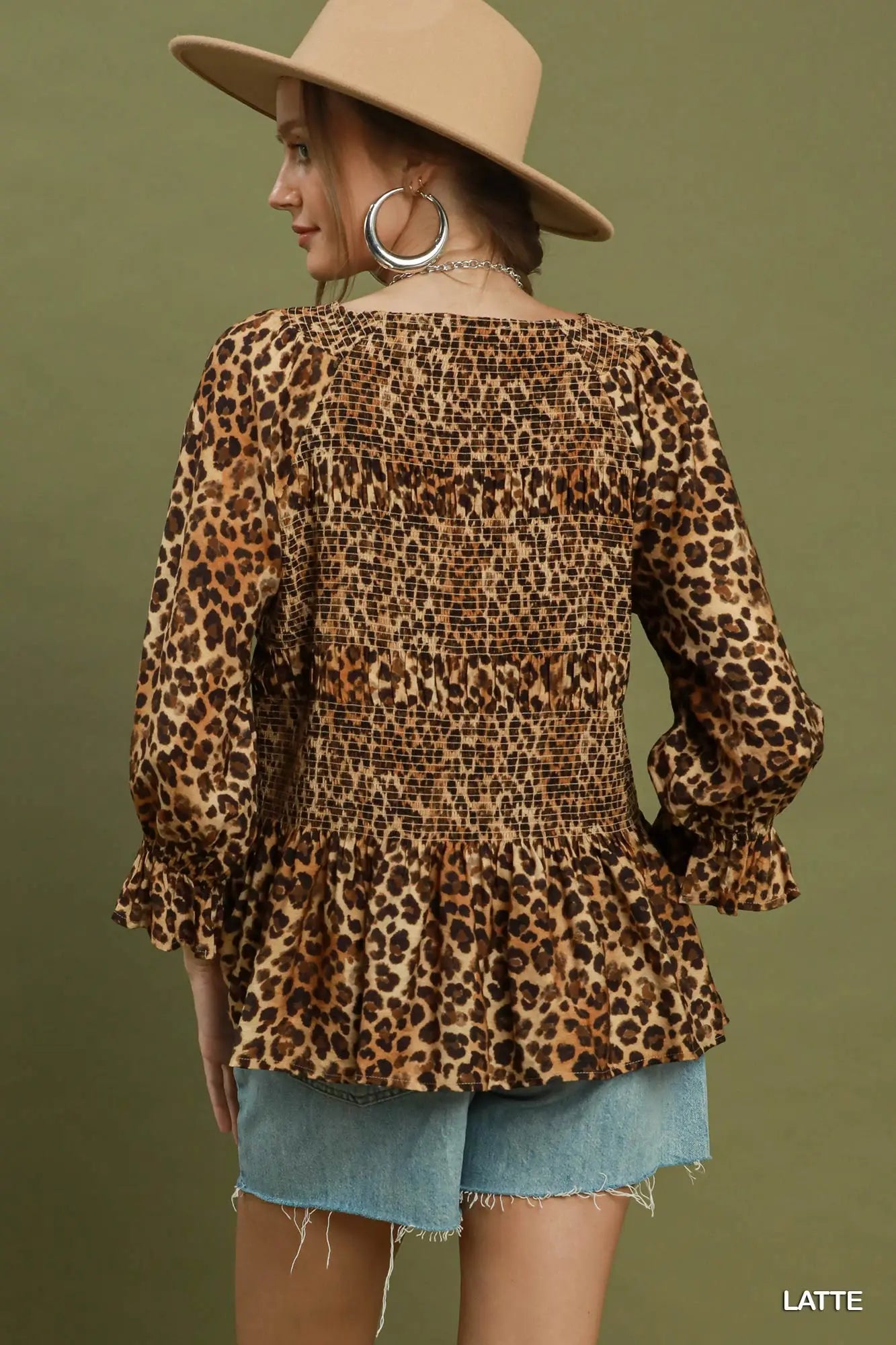 A woman wearing a leopard print smocked blouse with long balloon sleeves and a ruffled peplum hem, shown from the back against a solid olive green background.