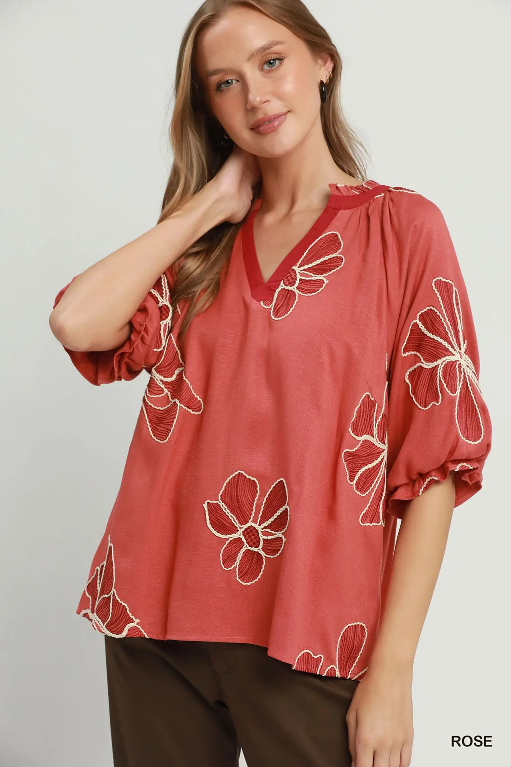 A woman wearing a loose, dusty rose V-neck blouse with white floral embroidery and puff sleeves, paired with dark brown pants, against a light gray studio background.