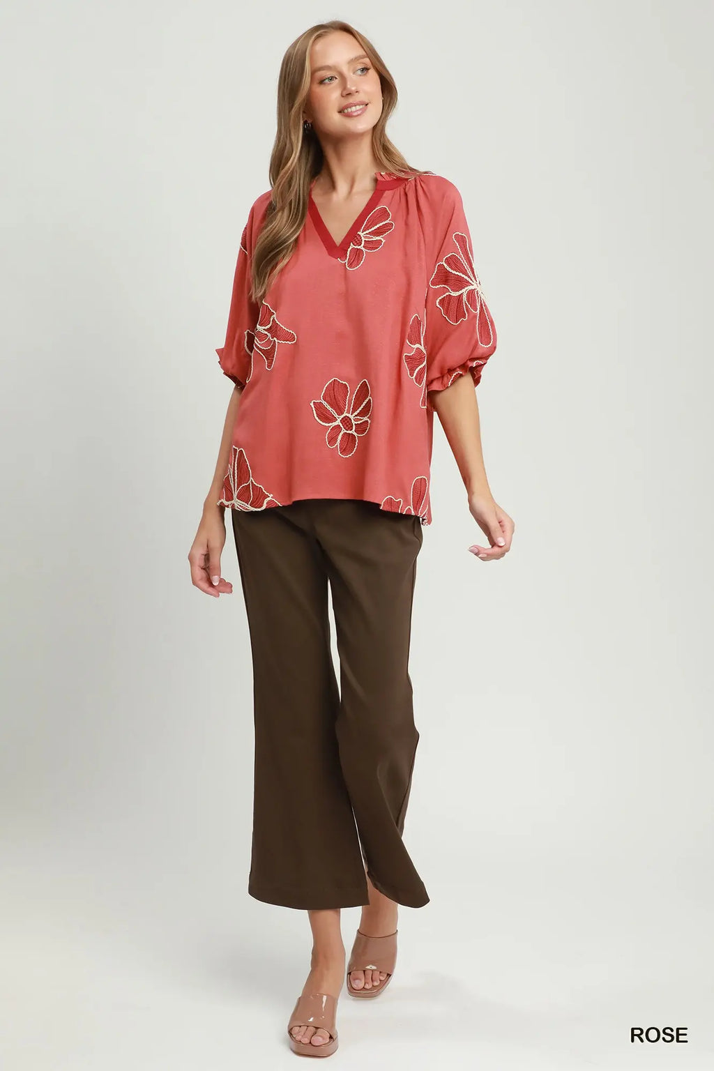A woman modeling a loose, short-sleeve blouse in dusty rose with white floral embroidery, paired with dark brown cropped pants, against a light gray studio background.