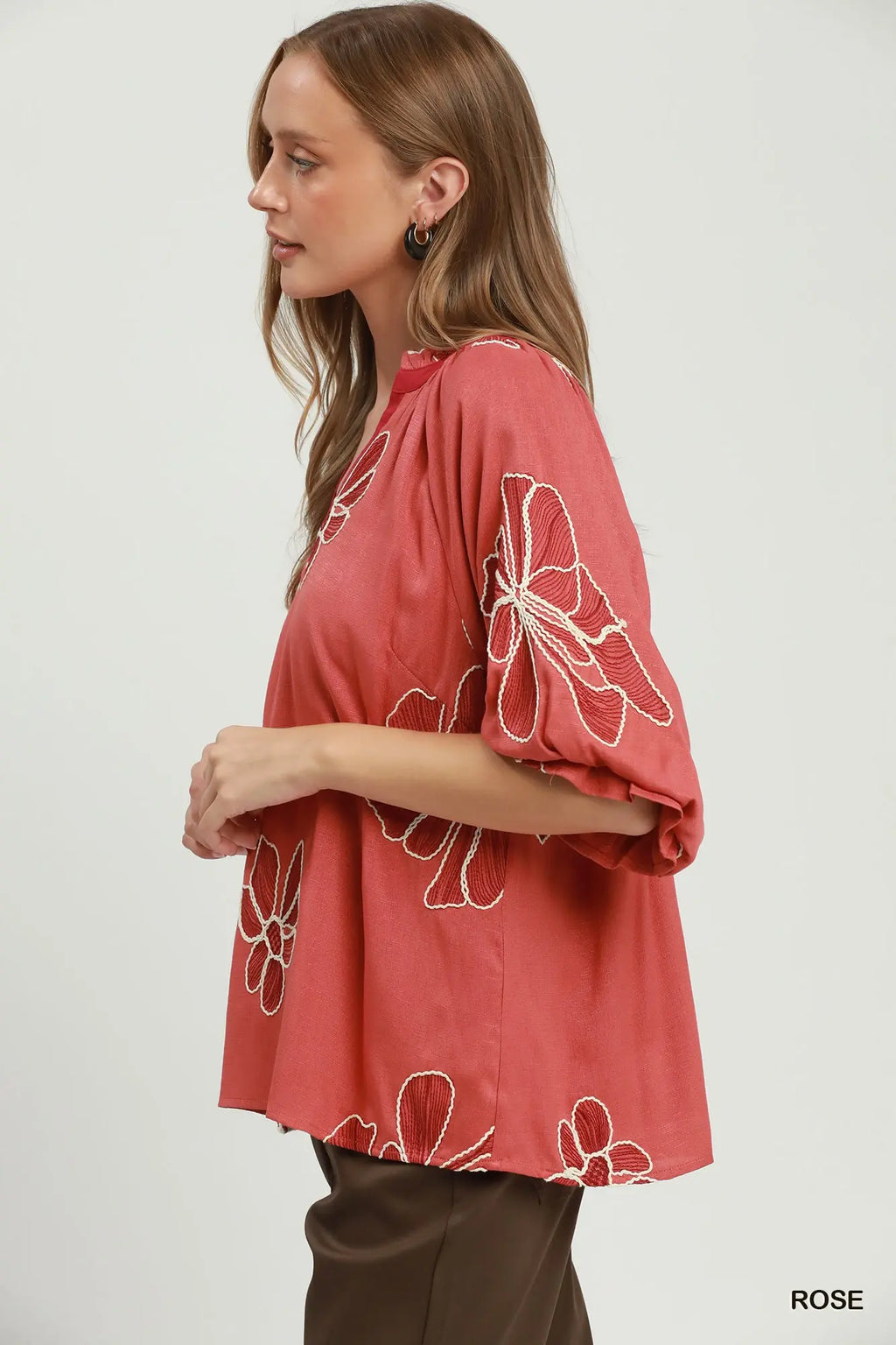 A woman wearing a loose, coral-rose short-sleeve top with white floral line embroidery, paired with brown pants, shown in profile against a light gray studio background.