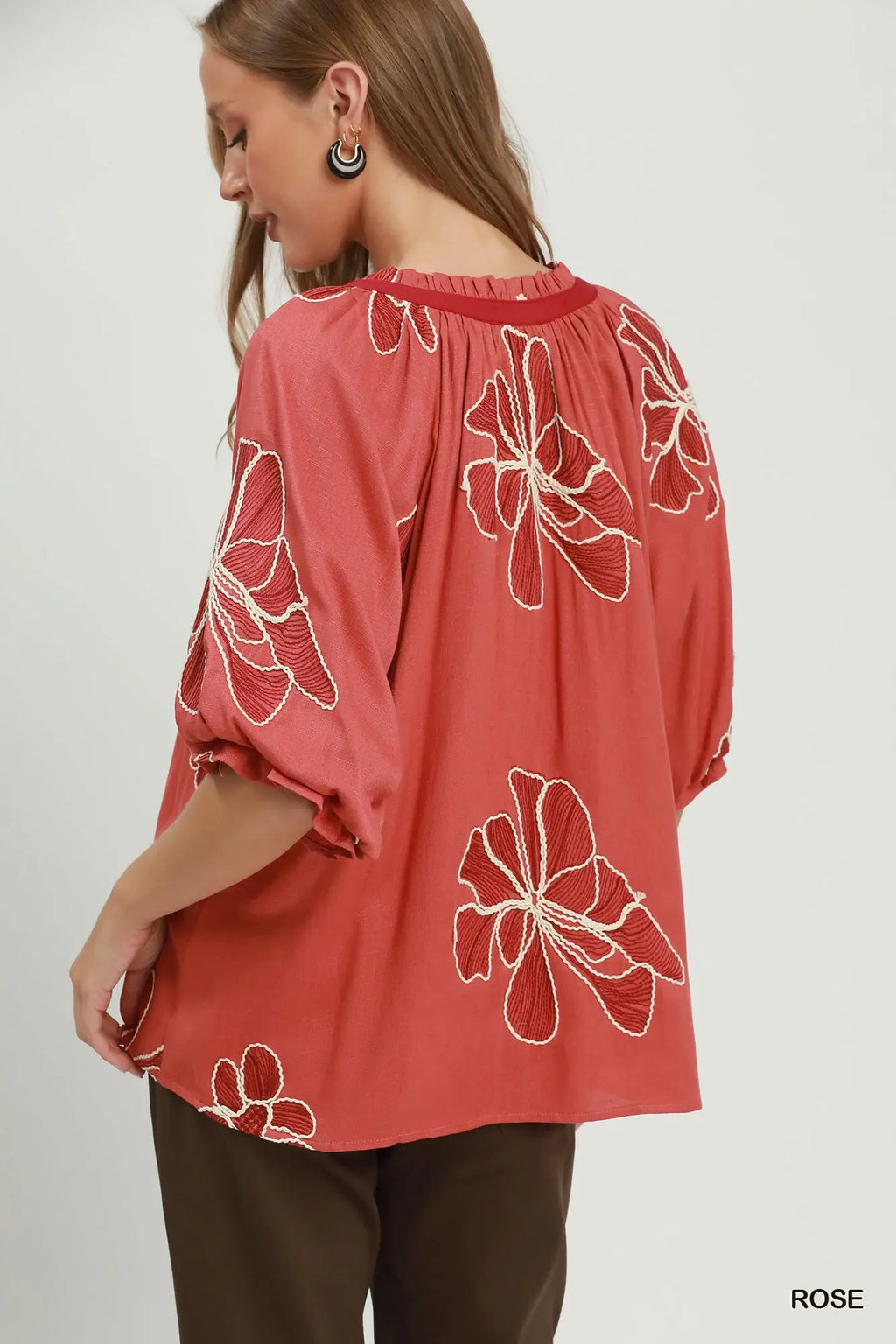A woman wearing a loose coral pink blouse with white floral embroidery, shown from the back against a light gray studio background. The word 'ROSE' appears in the bottom right corner.
