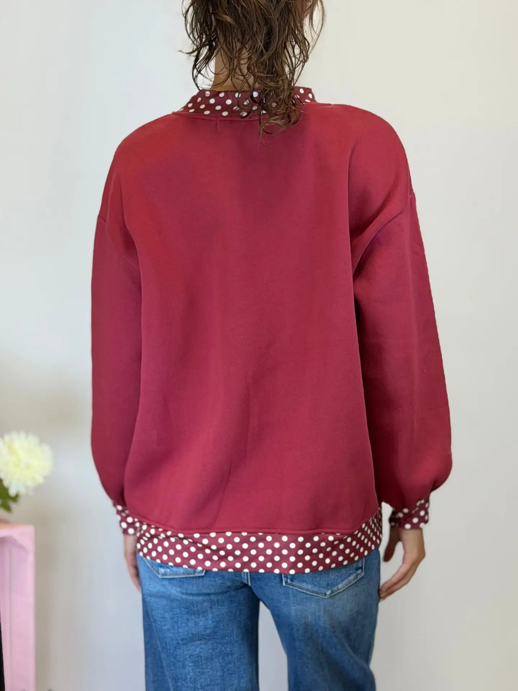 A person shown from the back wearing a dusty rose red long-sleeve sweatshirt with a contrasting red and white polka dot pattern on the collar and hem, paired with blue jeans.