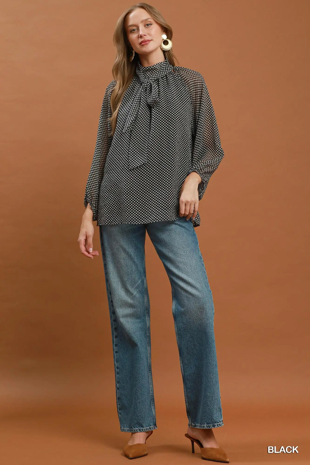 A woman modeling a black and white micro-patterned blouse with a tie-neck detail, paired with blue jeans and brown heels, against a solid brown studio background.