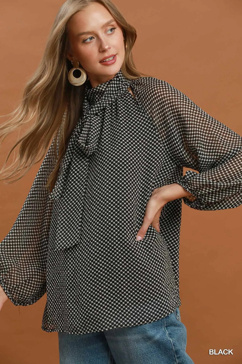 A woman wearing a black and white micro-check patterned blouse with a high neck and large bow detail, paired with blue jeans, set against a solid warm brown background.