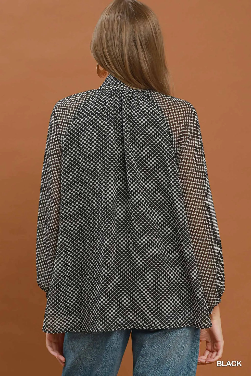 A woman shown from the back wearing a black and white micro-check patterned blouse with sheer mesh sleeves, paired with blue jeans, against a solid terracotta background.