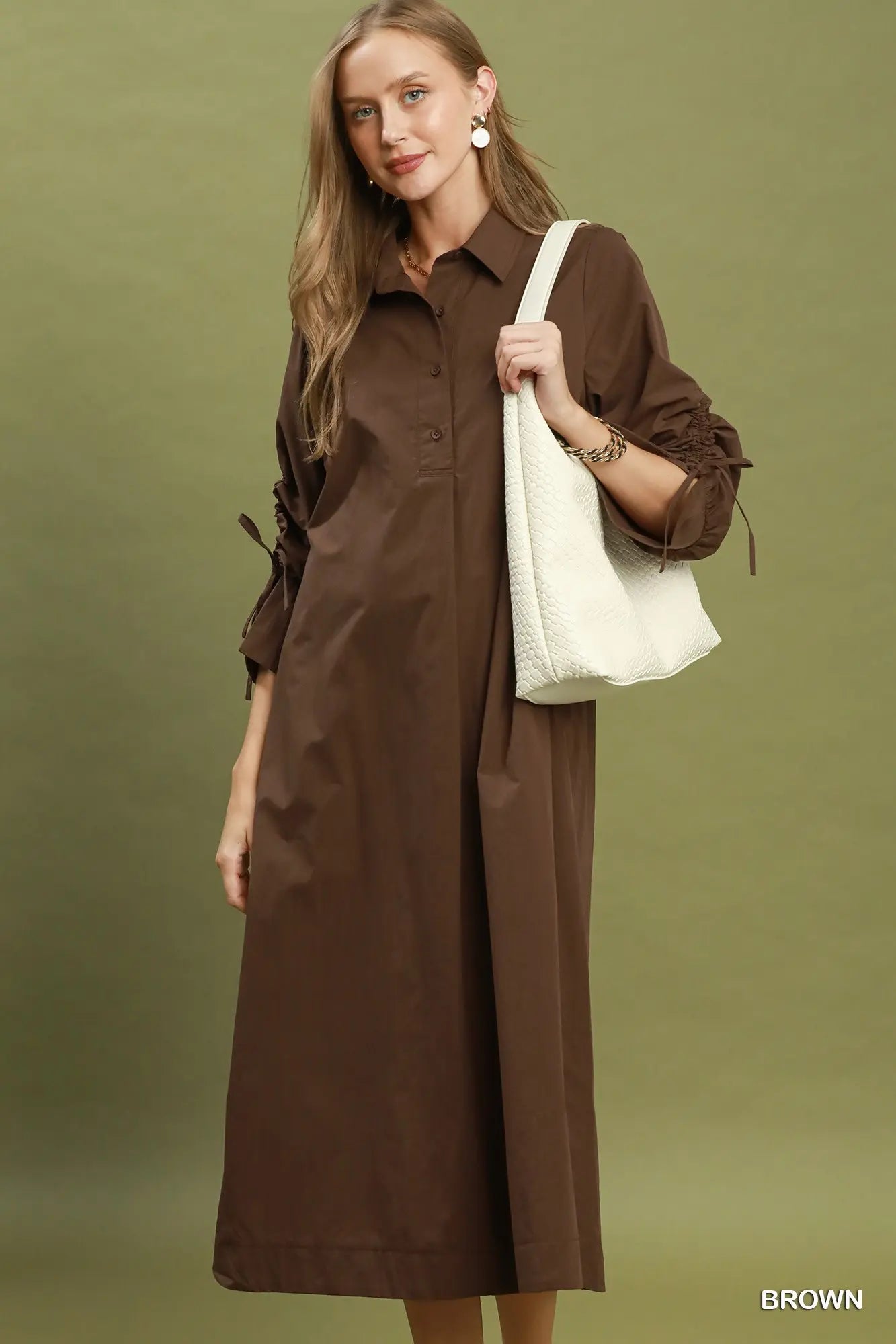 A woman modeling a brown long-sleeve button-front shirt dress with side tie details, carrying a white shoulder bag against an olive green studio background. The word 'BROWN' appears in the bottom right corner.