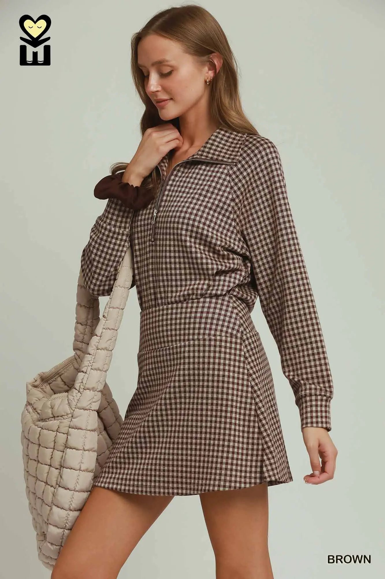 Woman wearing a brown checkered dress holding a beige quilted bag on a light gray background