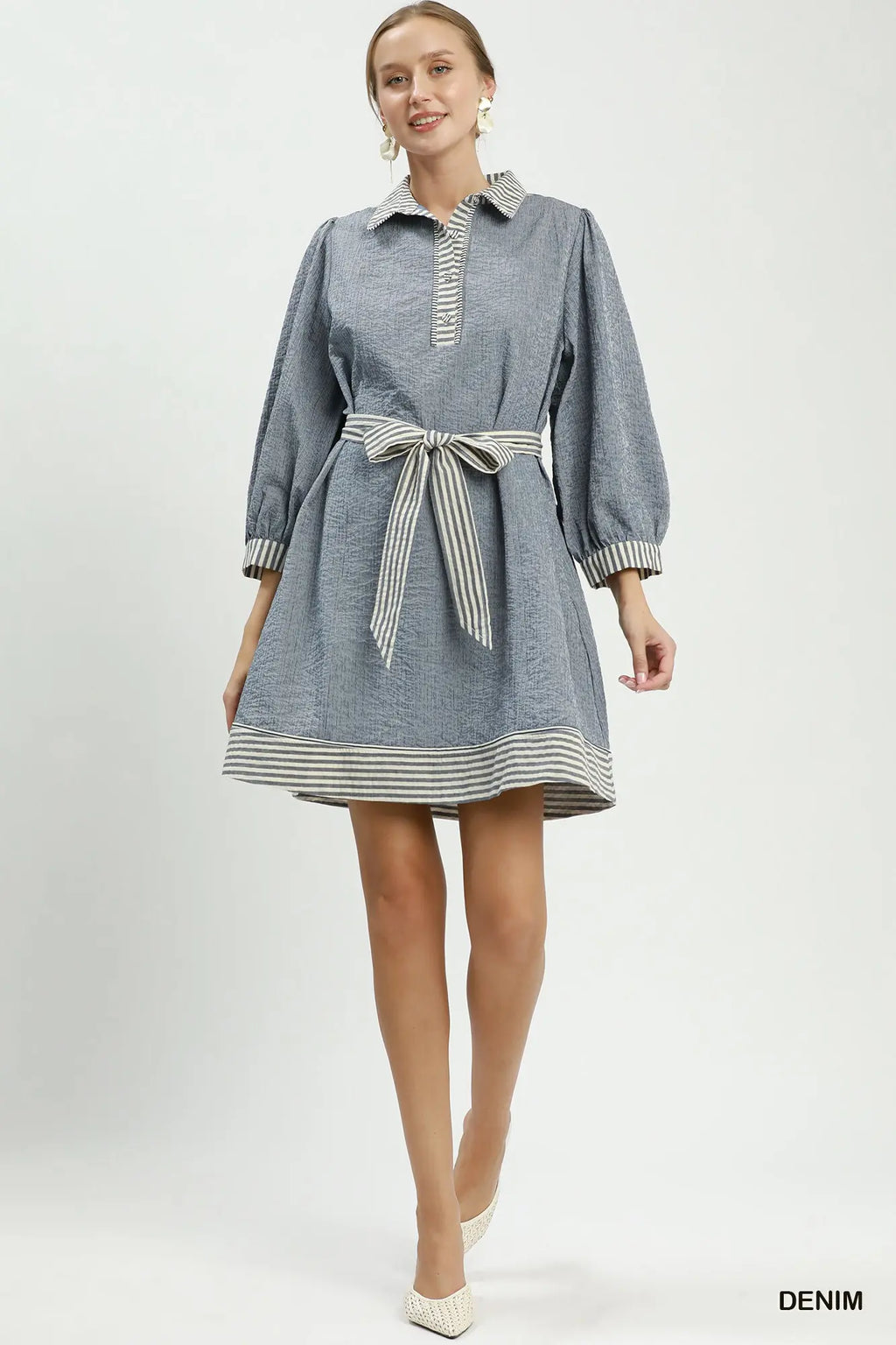 A woman modeling a light blue denim dress with striped trim on the collar, cuffs, hem, and matching striped tie belt, photographed against a plain light gray studio background.