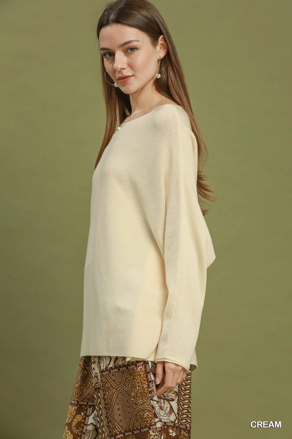 A woman wearing a cream-colored knit sweater and a brown patterned skirt, standing against a solid olive green background. The word 'CREAM' is printed in white text in the bottom right corner.