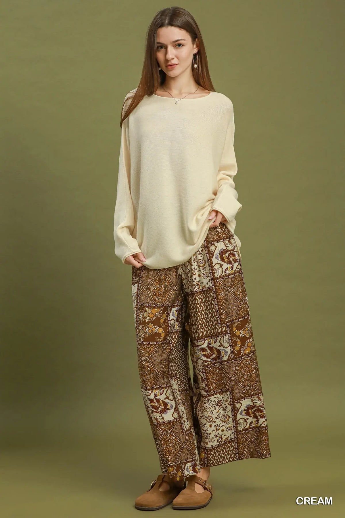 A woman modeling a cream long-sleeve knit top and brown patterned wide-leg pants against an olive green studio background. The word 'CREAM' appears in the bottom right corner.