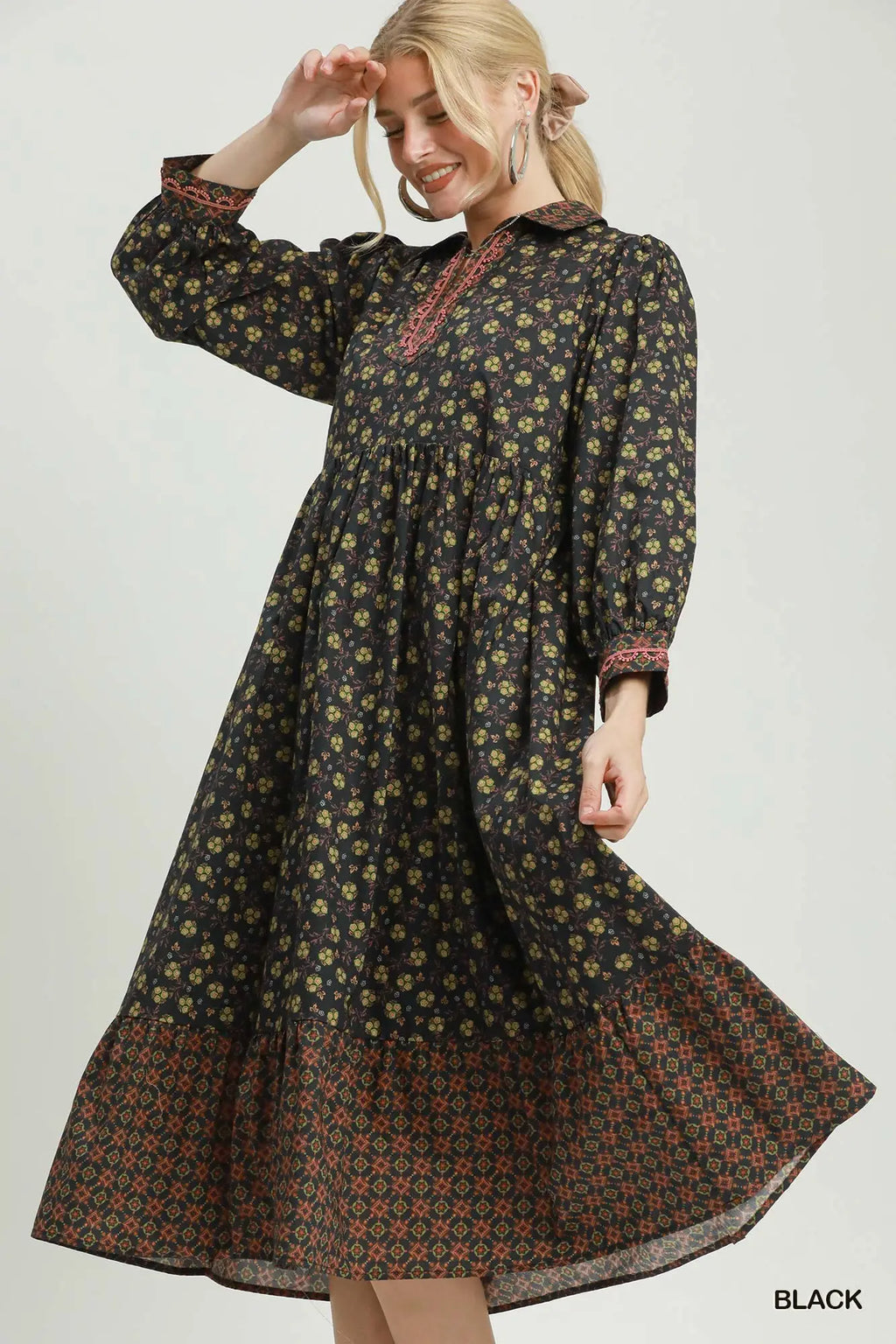 A woman modeling a long-sleeved black midi dress with a yellow and green floral print and a red patterned hem, set against a plain light gray background.