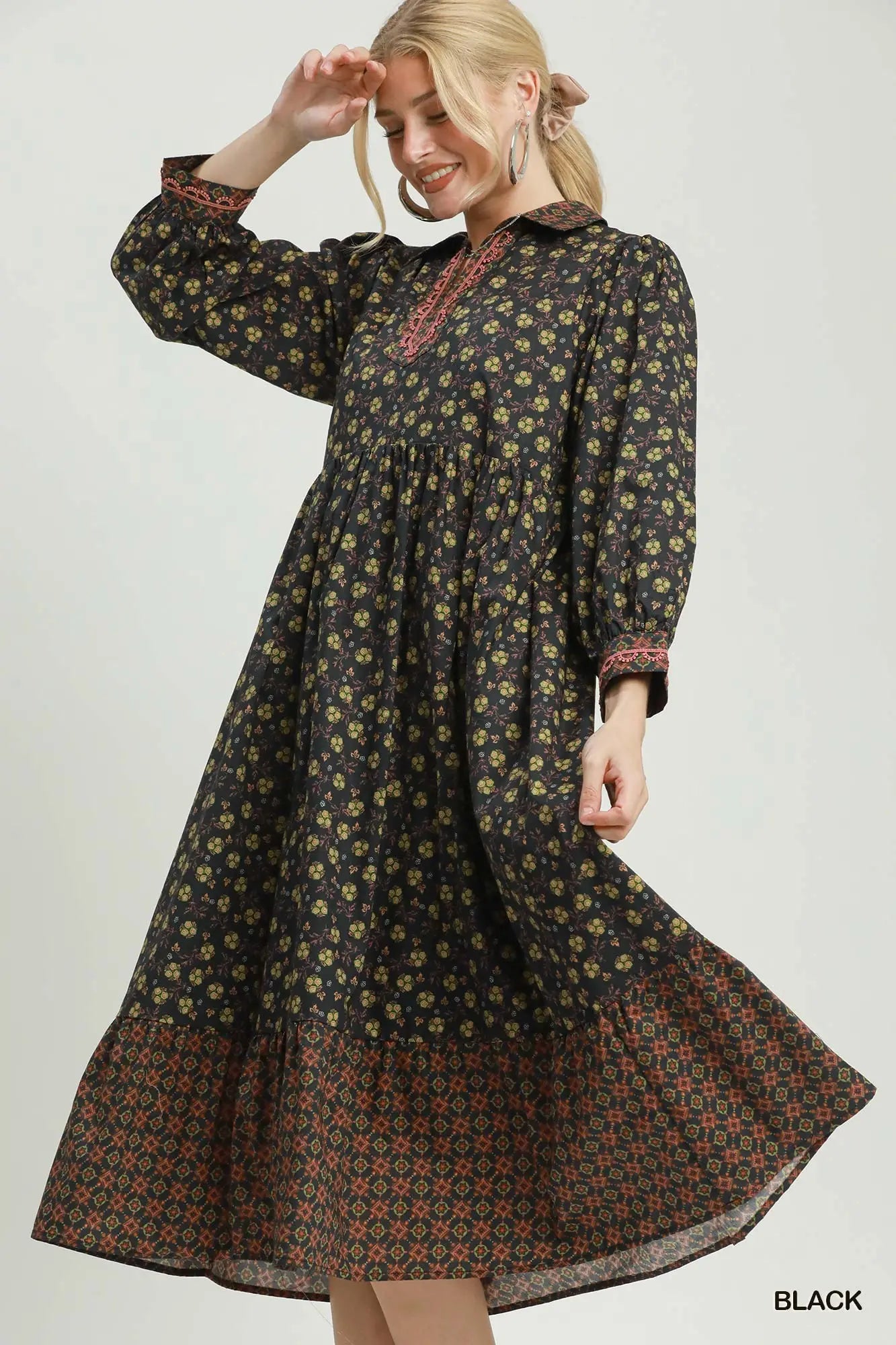 A woman modeling a long-sleeved black midi dress with a yellow and green floral print and a red patterned hem, set against a plain light gray background.