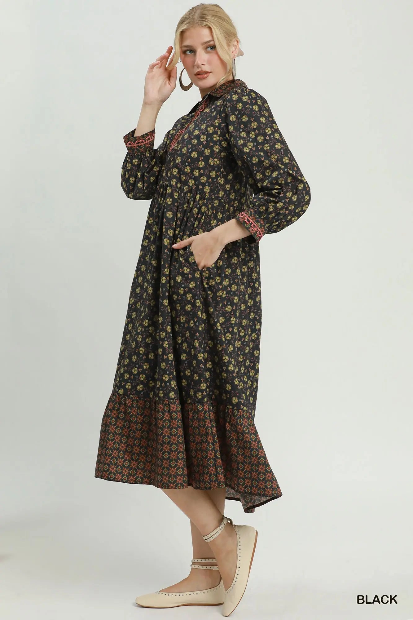 A woman modeling a long-sleeved black midi dress with a dense yellow and green floral print, featuring a contrasting patterned hem and cuffs, paired with beige pointed flats against a light gray studio background.