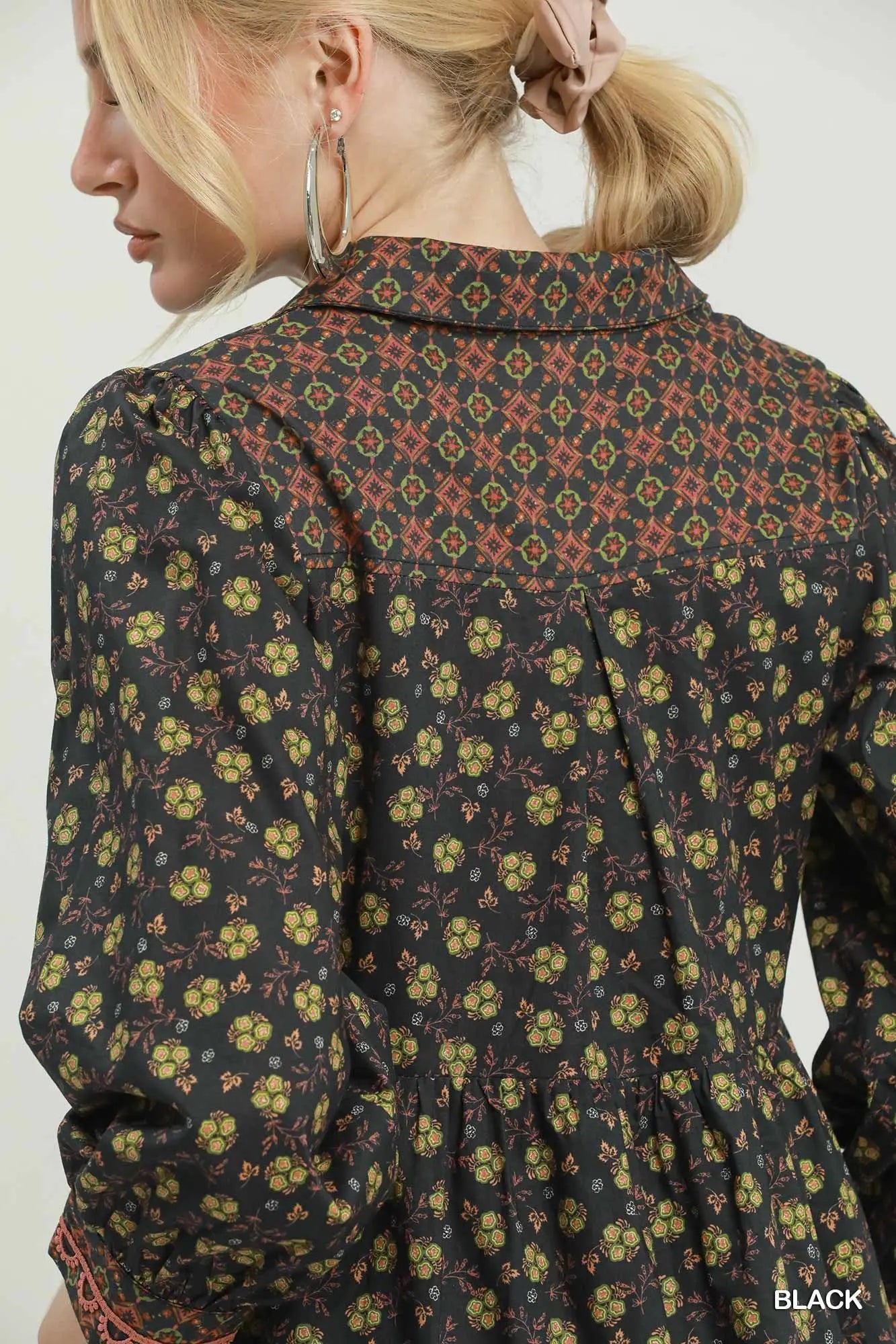 A woman shown from the back wearing a long-sleeve collared blouse with a dense floral and geometric print on a black base. The print features yellow, orange, and red motifs.