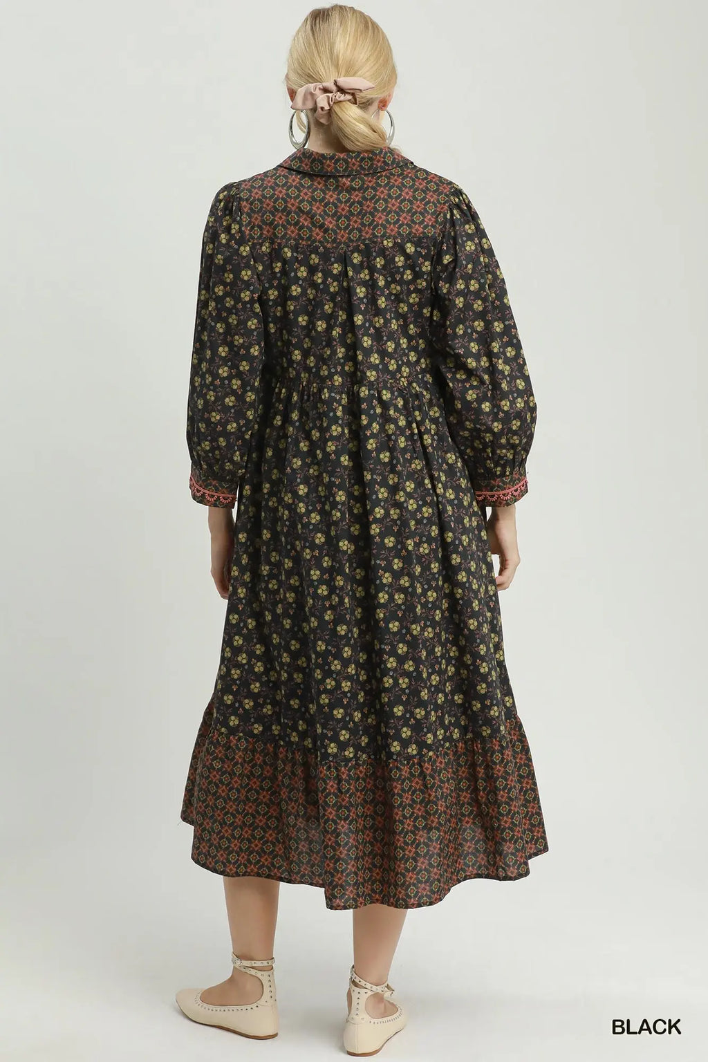 A woman shown from the back wearing a long-sleeved midi dress with a black base and a dense small floral print in muted yellow, green, and rust tones. The dress features a high collar, gathered waist, and a tiered ruffle hem.