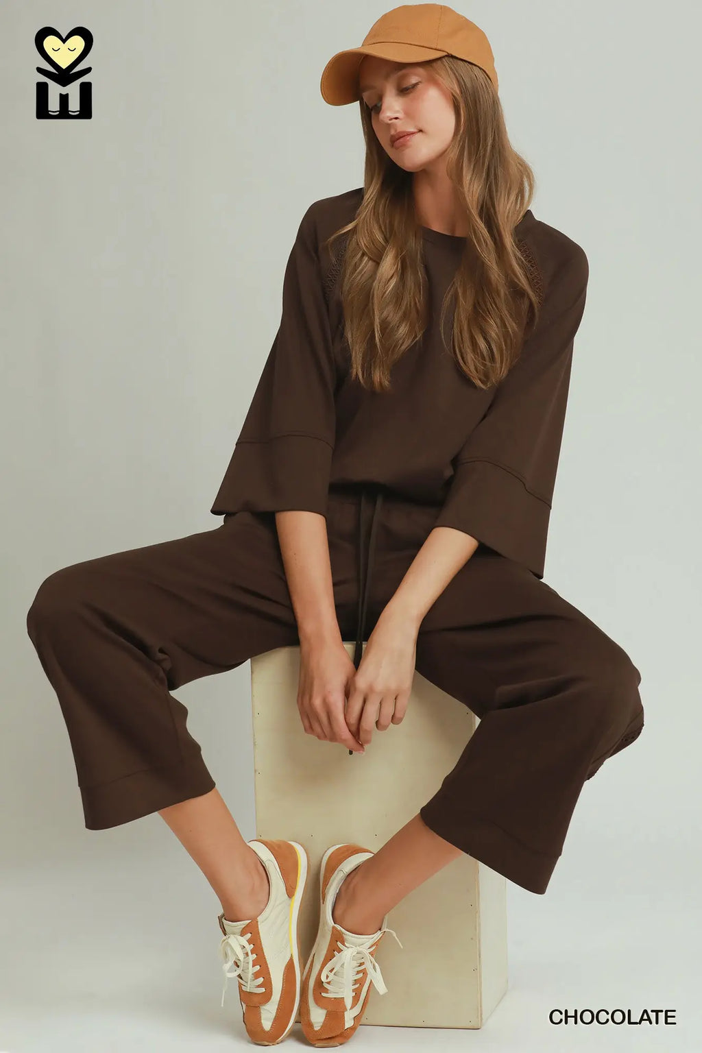 Woman in brown outfit with beige cap and sneakers sitting on a box against a white background