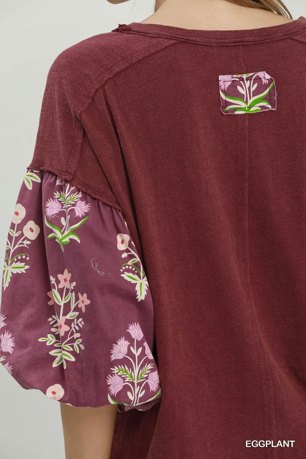 A close-up of a person wearing a burgundy top with floral-patterned puff sleeves and a small floral patch on the upper back. The text 'EGGPLANT' appears in the bottom right corner.