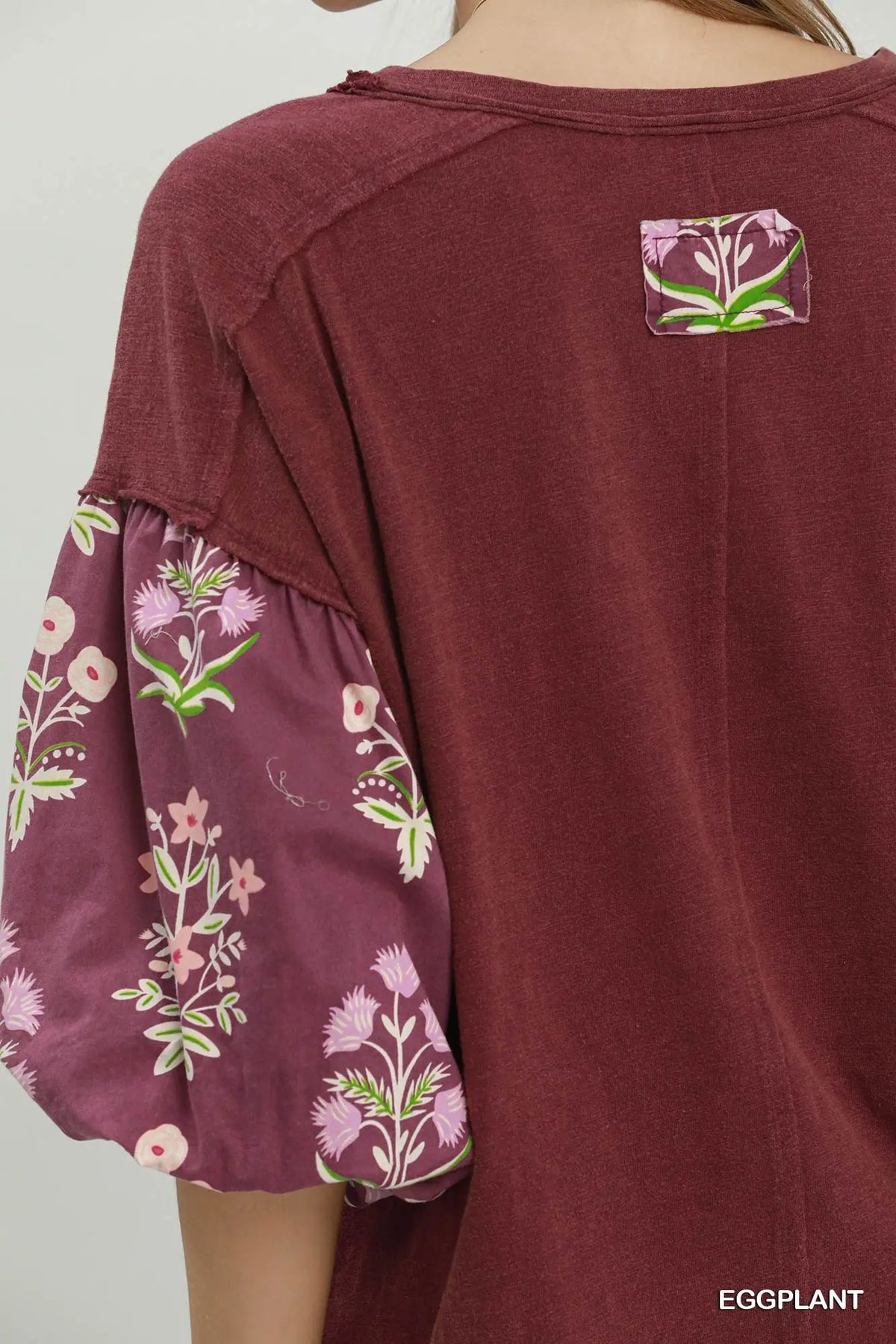 A close-up of a person wearing a burgundy top with floral-patterned puff sleeves and a small floral patch on the upper back. The text 'EGGPLANT' appears in the bottom right corner.