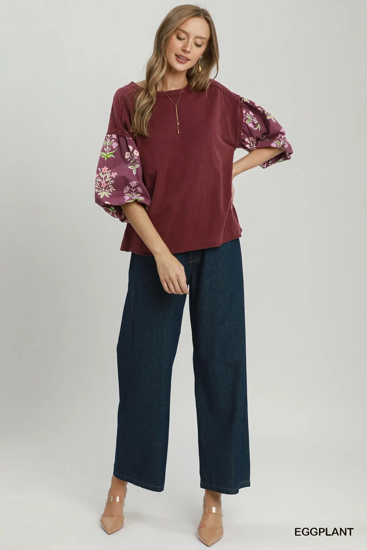 A woman modeling a burgundy top with floral-patterned puff sleeves and dark wide-leg jeans, standing against a light gray studio background. The word 'EGGPLANT' appears in the bottom right corner.