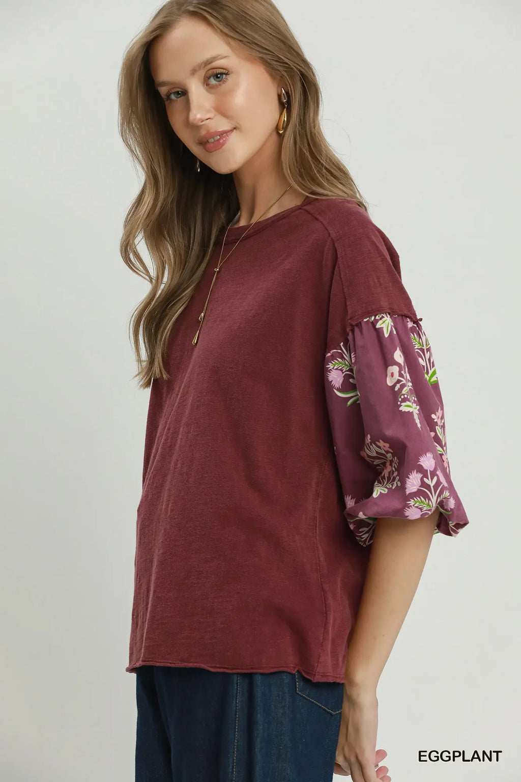 A woman wearing a burgundy top with floral-patterned puff sleeves, paired with dark blue jeans, photographed against a light gray studio background. The brand name 'EGGPLANT' is visible in the bottom right corner.