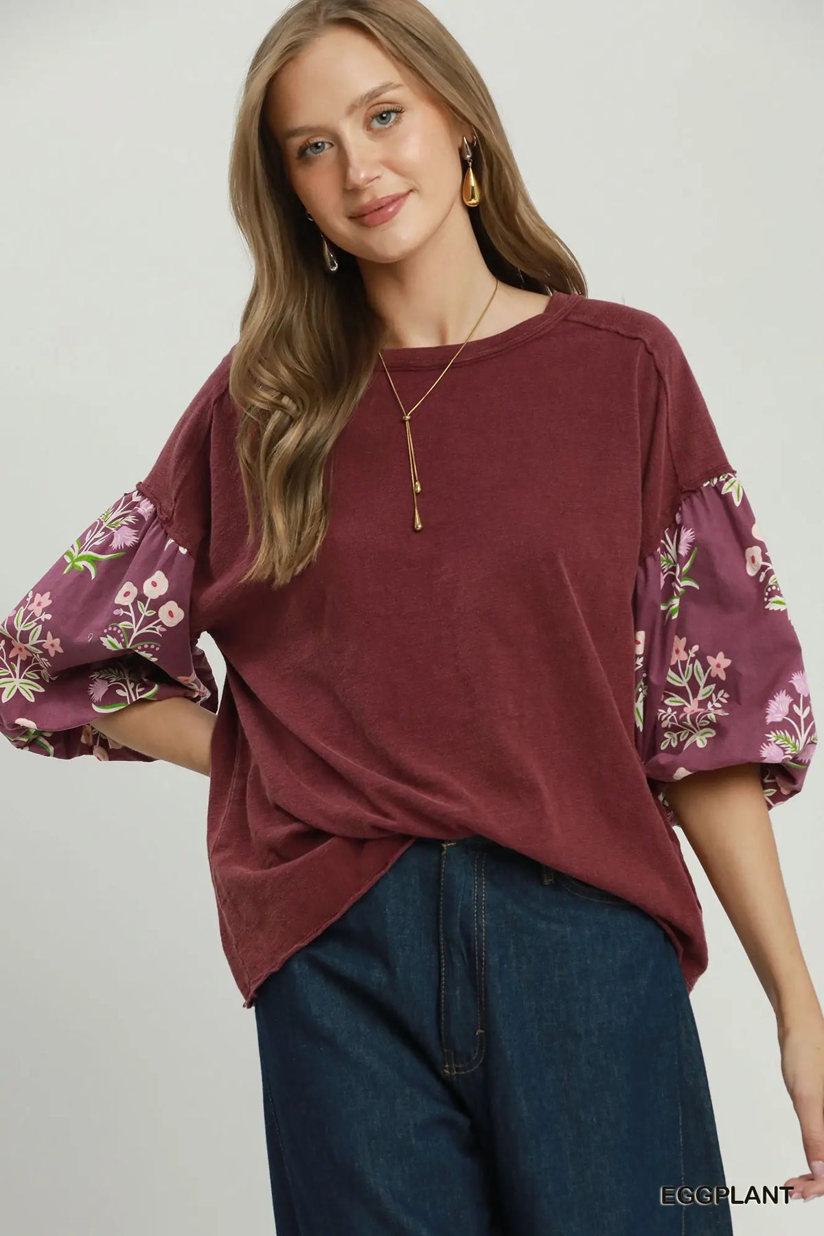 A woman wearing a loose-fitting dusty rose top with floral puff sleeves and dark blue jeans, photographed against a plain light gray studio background. The brand name EGGPLANT is visible in the bottom right corner.