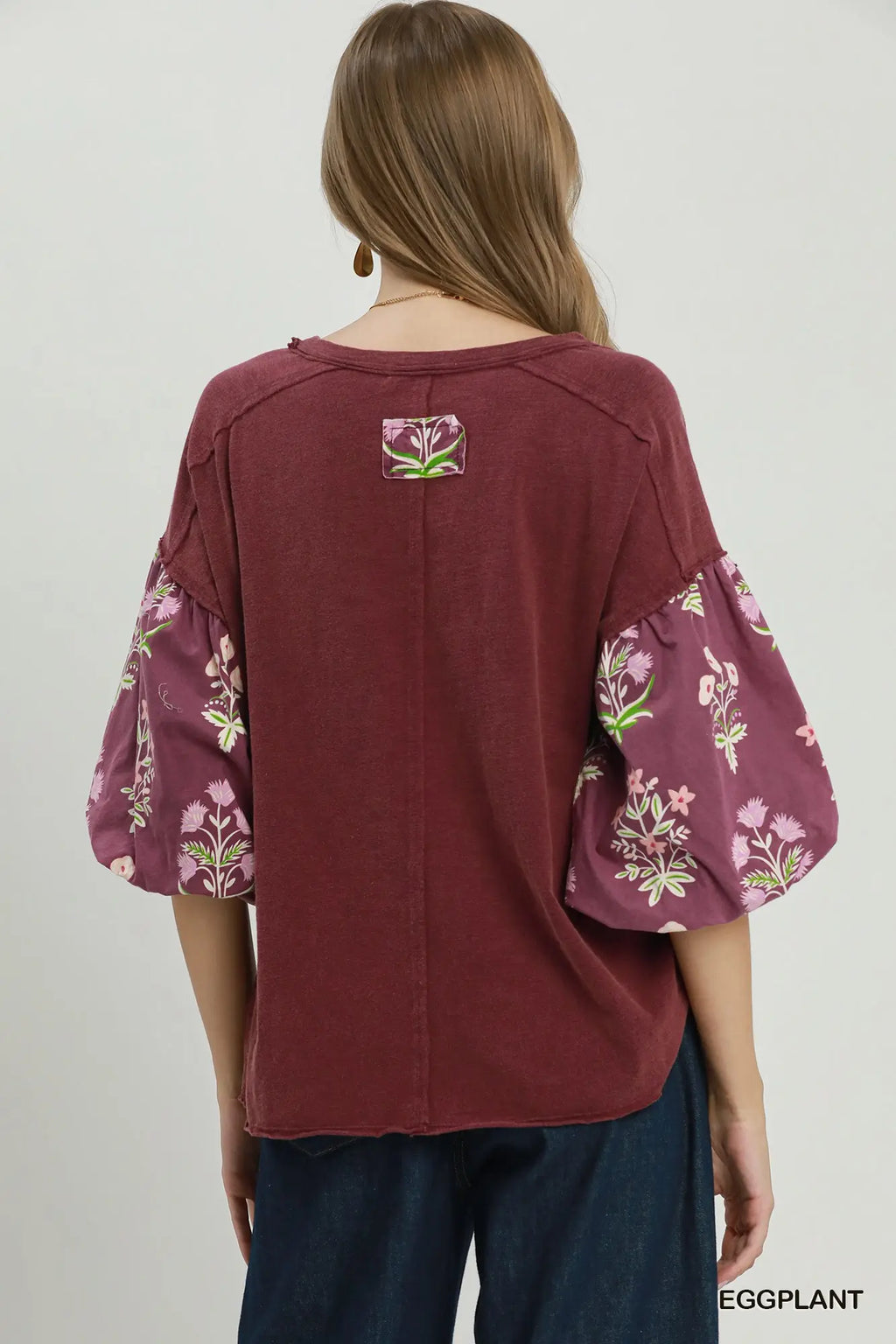 A woman shown from the back wearing a burgundy top with floral-patterned puff sleeves and dark blue jeans, against a light gray studio background.