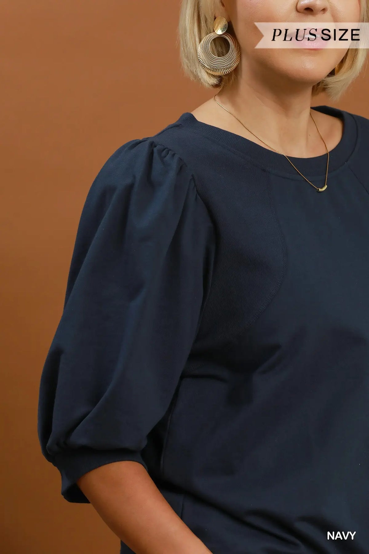 A close-up of a woman wearing a navy blue blouse with voluminous elbow-length puff sleeves and a round neckline, set against a solid warm brown background. Text overlays read 'PLUSSIZE' and 'NAVY'.