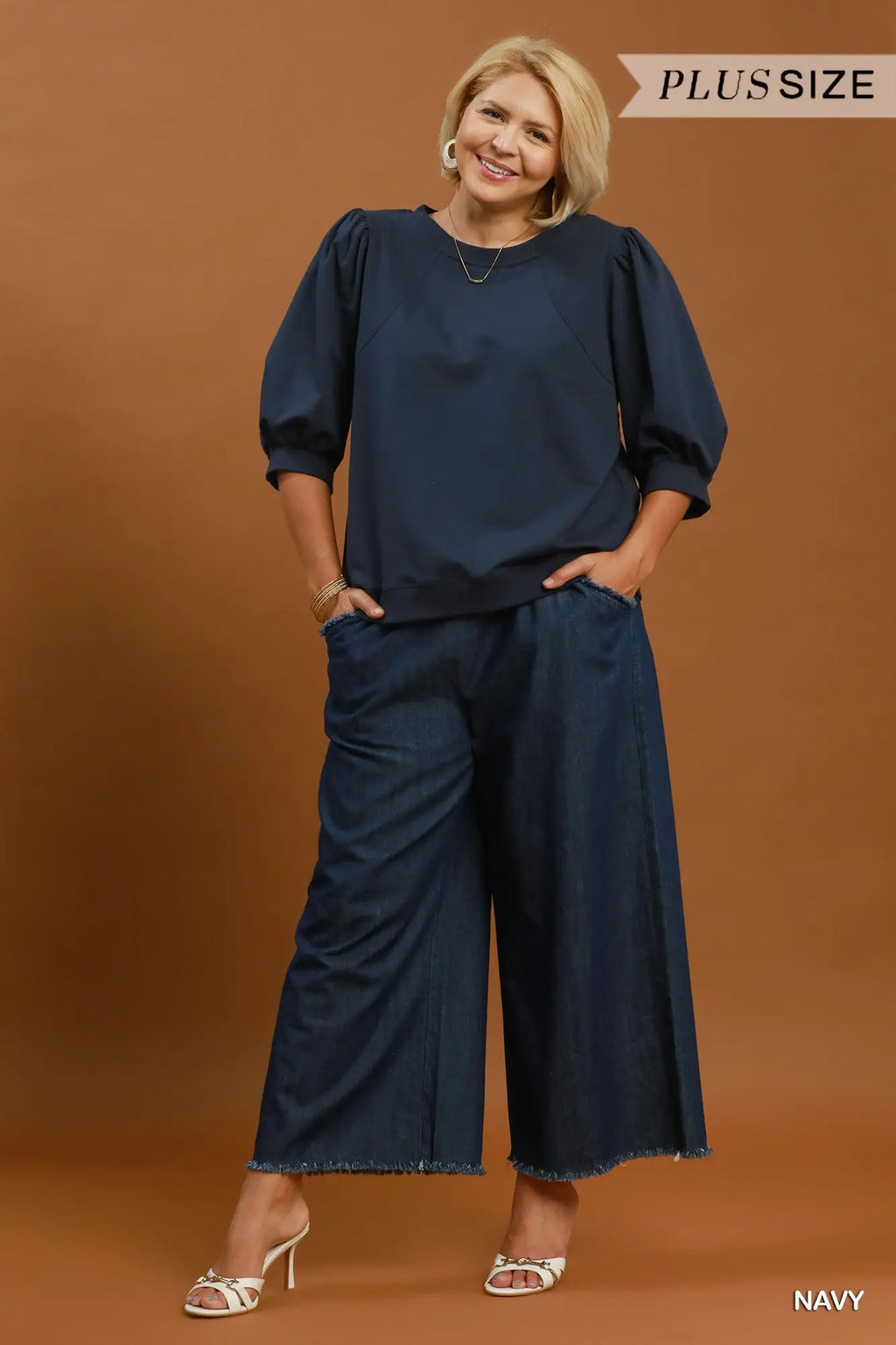 A woman modeling a navy blue two-piece outfit consisting of a long-sleeve top with puff sleeves and matching wide-leg pants, set against a solid brown studio background. Text overlays read 'PLUS SIZE' at the top right and 'NAVY' at the bottom right.
