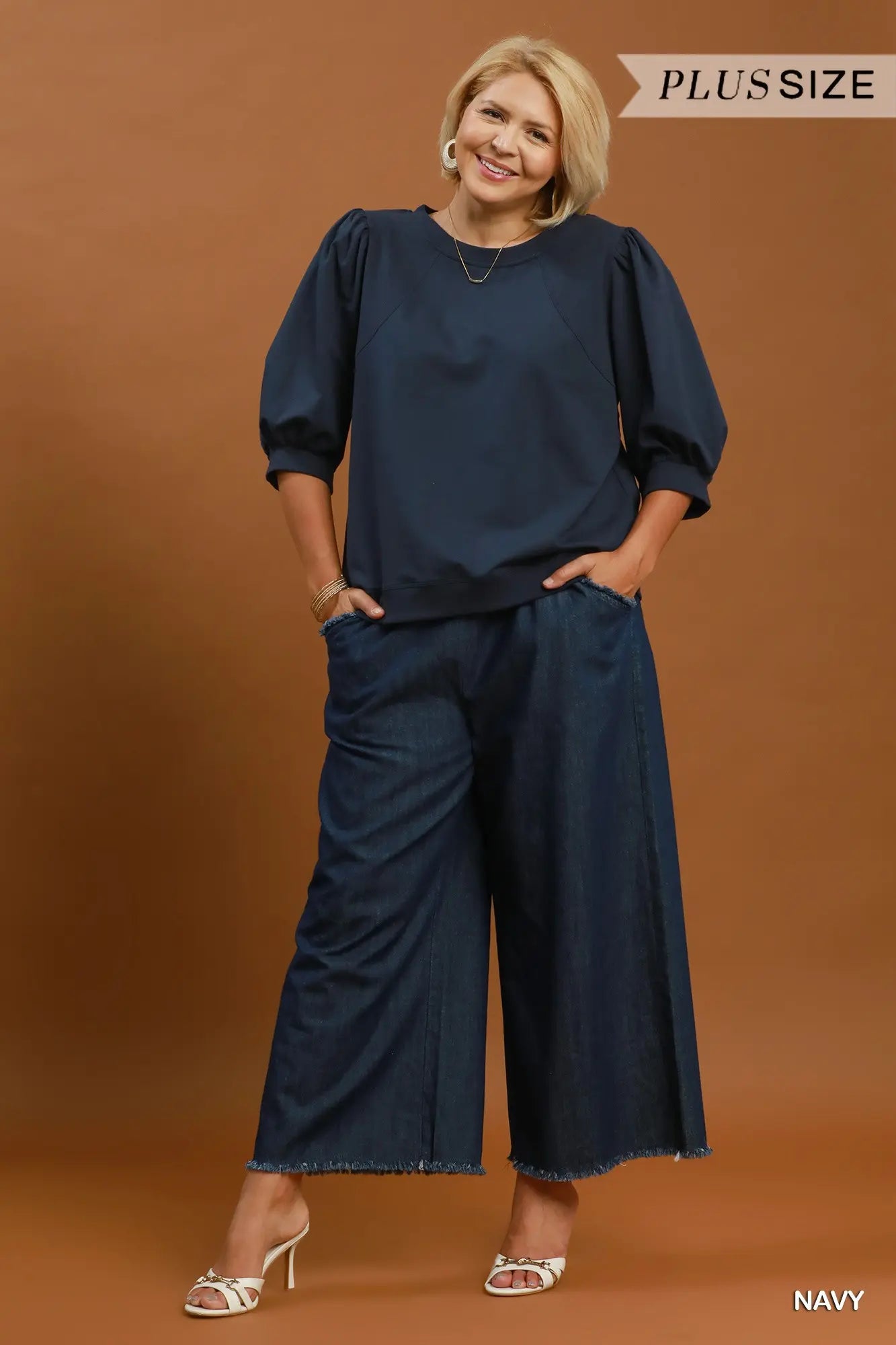 A woman modeling a navy blue two-piece outfit consisting of a long-sleeve top with puff sleeves and matching wide-leg pants, set against a solid brown studio background. Text overlays read 'PLUS SIZE' at the top right and 'NAVY' at the bottom right.