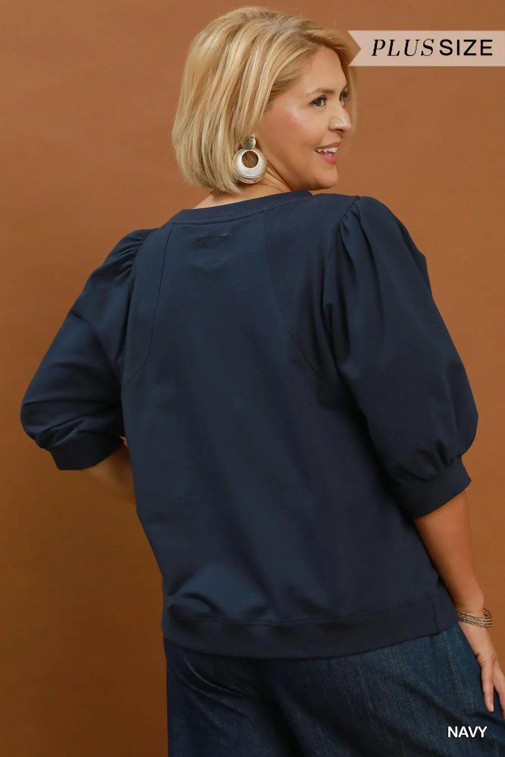 A woman modeling a navy blue puff-sleeve top from the back, shown against a solid warm brown studio background. The image includes 'PLUS SIZE' text in the top right and 'NAVY' text in the bottom right.