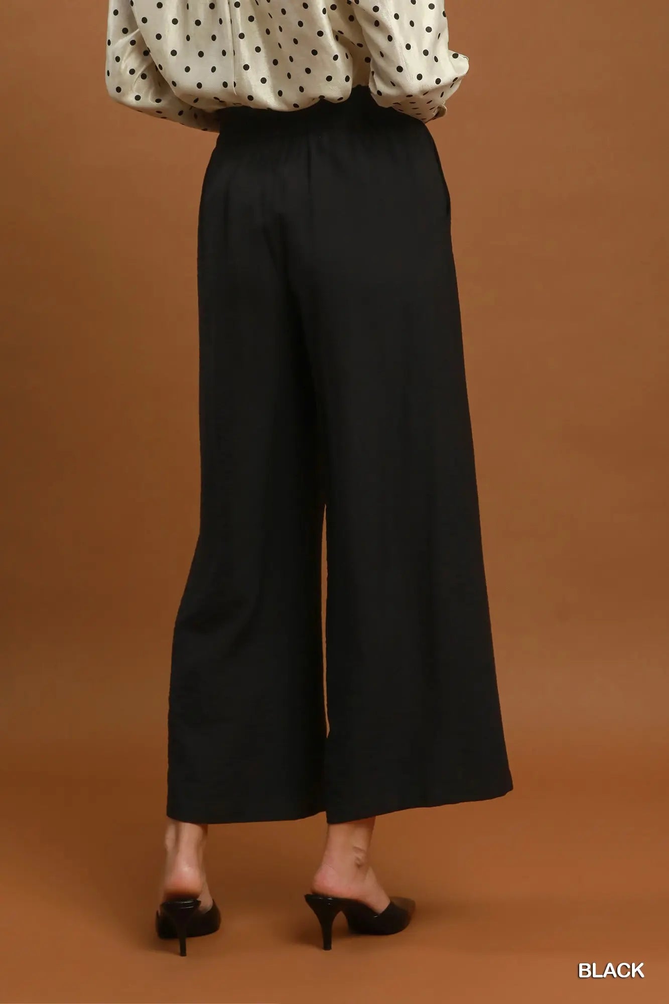 A model is shown from the back wearing black wide-leg cropped pants, a cream blouse with black polka dots, and black pointed-toe mule heels against a solid warm brown studio background. The word 'BLACK' is printed in white in the bottom right corner.