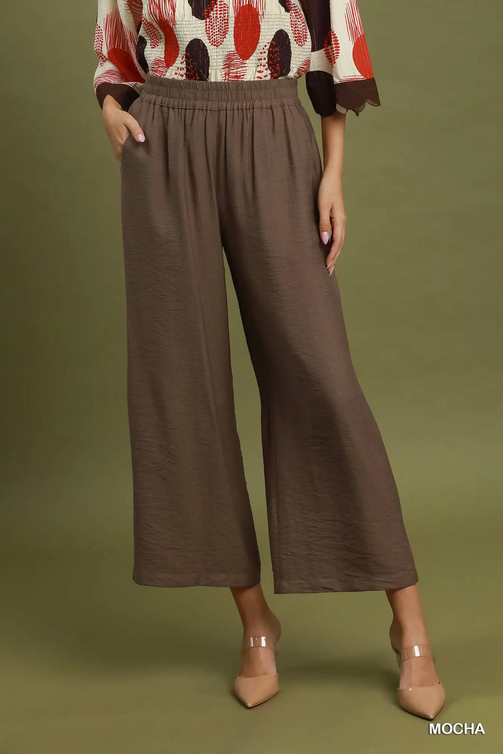A model wearing a pair of wide-leg cropped trousers in a solid mocha brown color, paired with a patterned top and nude heels, set against a solid olive green studio background.