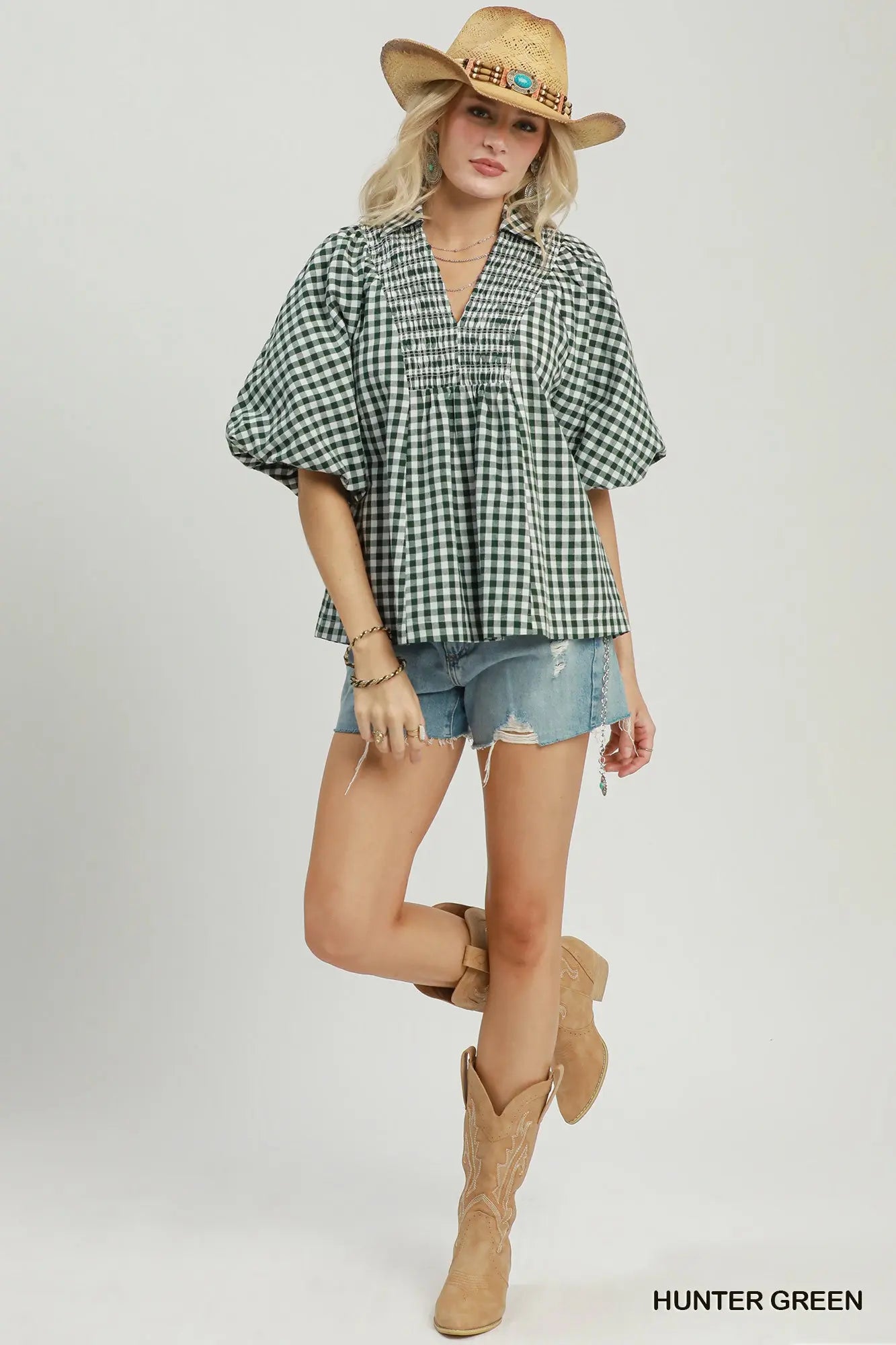 A woman modeling a green and black gingham short-sleeve blouse with puff sleeves, paired with denim shorts and tan cowboy boots, against a light gray studio background.