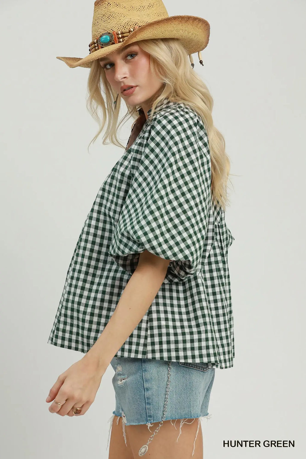 A woman wearing a green and white gingham short-sleeve shirt, denim shorts, and a straw cowboy hat with turquoise accents, posed against a light gray studio background. The text 'HUNTER GREEN' appears in the bottom right corner.