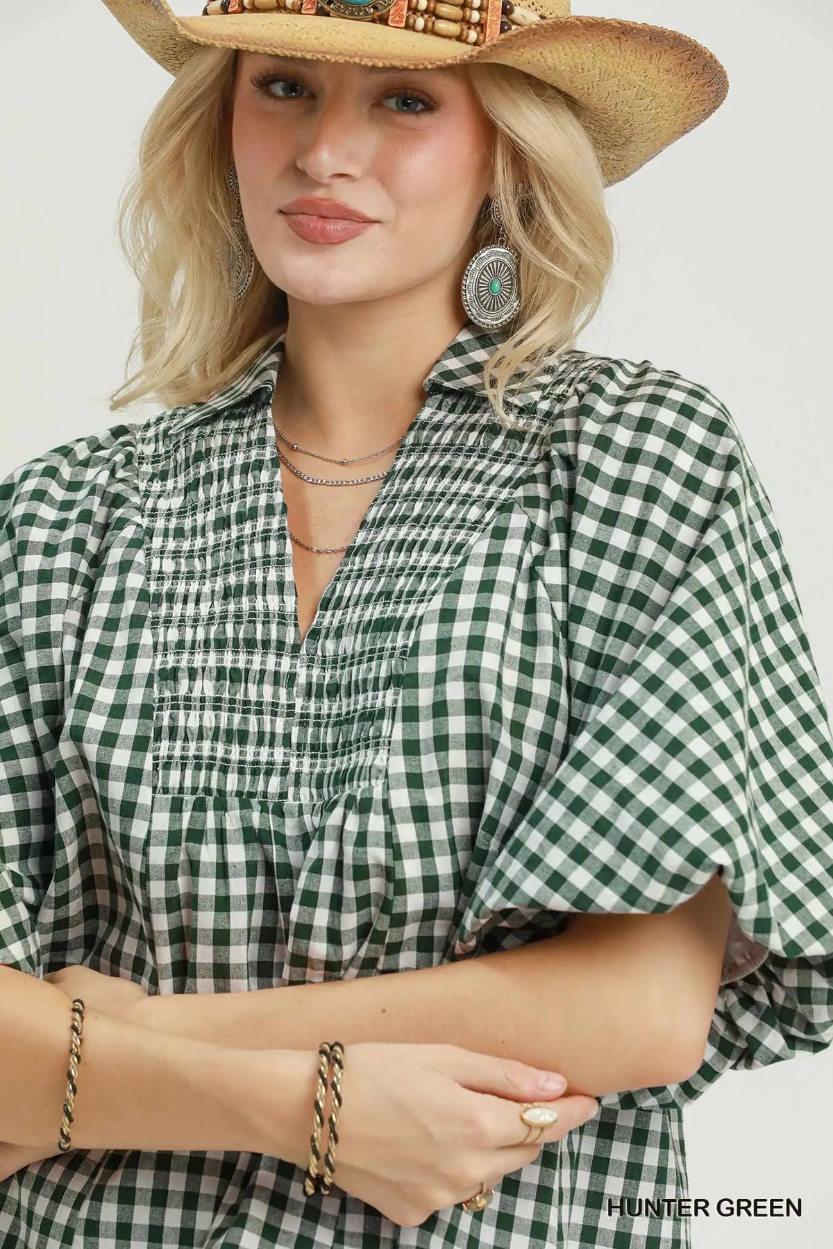 A woman wearing a green and white gingham blouse with a smocked chest and ruffled short sleeves, styled with a straw cowboy hat, layered necklaces, and bracelets against a light gray studio background.